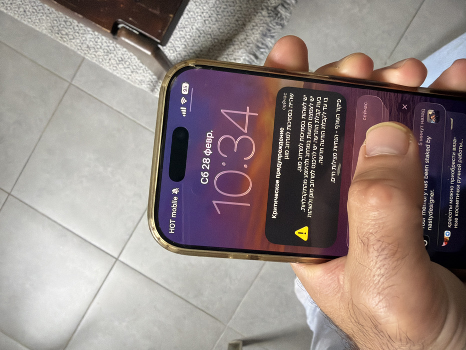 An adult male's right hand, with visible hair on the back, holds an iPhone in a golden/champagne-colored protective case. The thumb is positioned over the phone screen, near an interactive element of a critical alert. The phone screen displays the time "10:34" and date "Сб 28 февр." (Saturday, February 28th) in Cyrillic script. The carrier is "HOT mobile" and the battery level is 95%. A prominent black notification box with a yellow triangular warning icon occupies the central part of the screen. The notification title is "Критическое предупреждение" (Critical warning) in Russian. The body of the alert is primarily in Hebrew, stating "יש לשהות בסמיכות למרחב מוגן" (stay near a protected area) and providing further instructions from "פיקוד העורף - הנחיות מצילות חיים." (Home Front Command - Life-saving instructions). Below this, other notifications are partially visible, including one from "nastyldesigner". The background consists of light-colored, square floor tiles arranged diagonally, with a section of a patterned rug and a dark wooden object (possibly furniture) visible on the right. The presence of a "HOT mobile" network indication and an alert from the "Home Front Command" in Hebrew definitively places the scene within Israel, specifically consistent with an urban setting like Tel-Aviv.