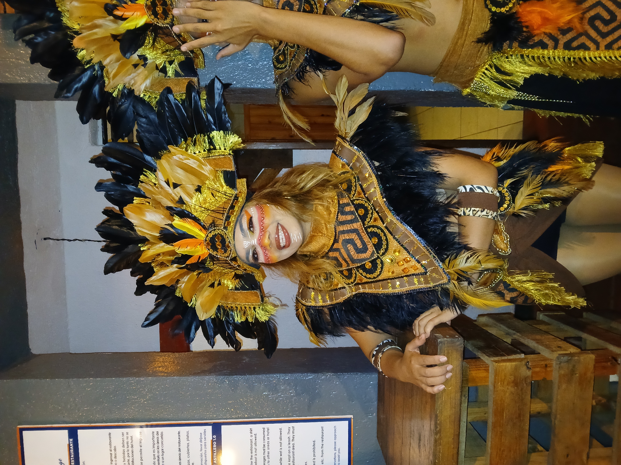 The image shows a woman wearing a black and gold feathered headdress and a matching decorative collar. Her face is painted with geometric patterns. She's leaning on a wooden structure, possibly a pallet. Another person wearing similar attire is partially visible in the upper right corner. The background includes a wall with a partially visible menu. The text on the menu appears to be in both Spanish and English, mentioning restaurant rules, such as dress code and beverage consumption policies. The location is potentially a restaurant in Porlamar, Venezuela.