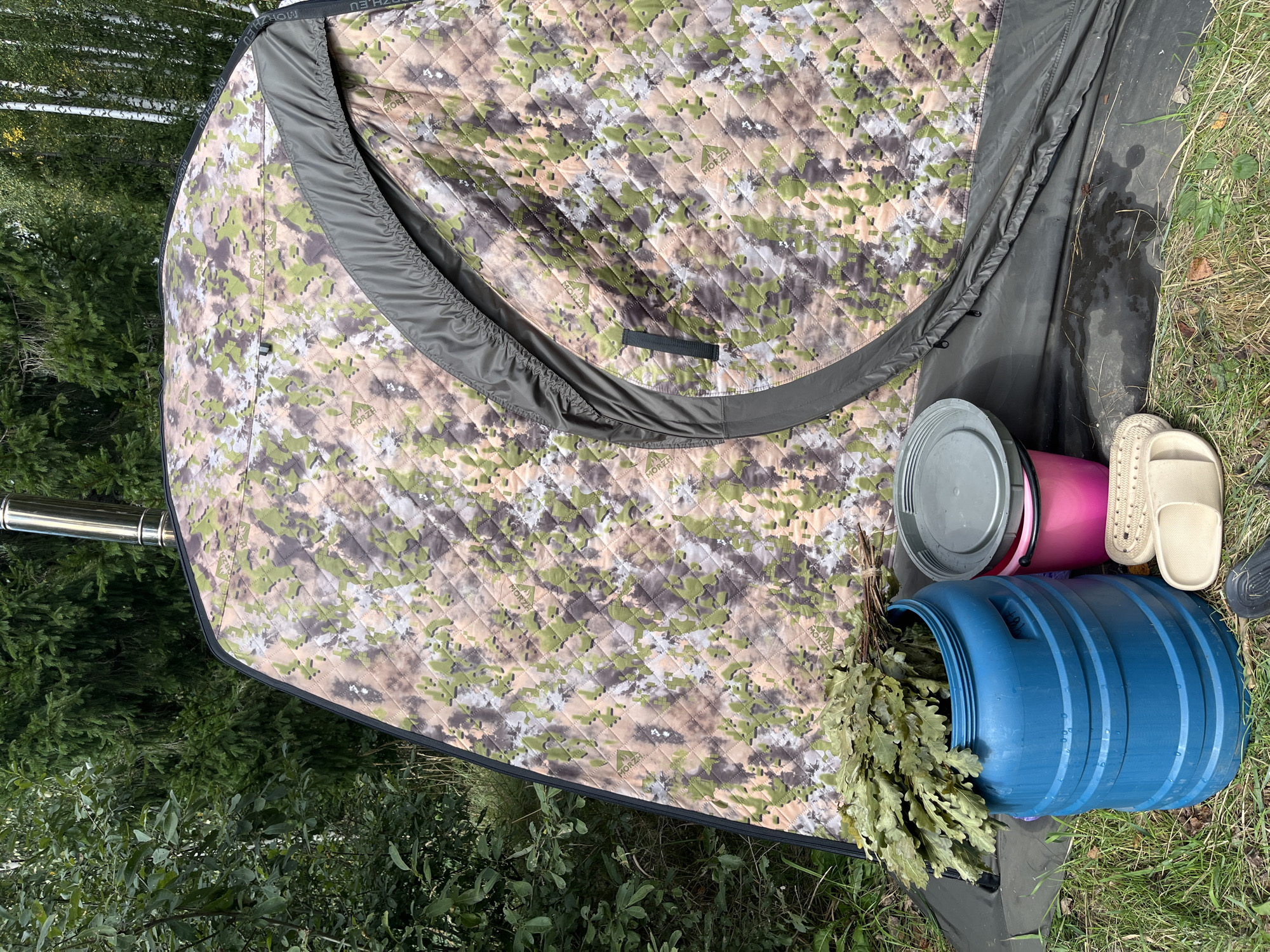 The image shows an outdoor scene in Novyi, Russia, with a camouflage-patterned portable sauna tent as the central subject. To the left of the tent, there is a large blue plastic barrel filled with green leafy branches, likely for a banya (Russian sauna). In front of the barrel, there are a pink bucket with a gray lid and a pair of beige flip-flops. The ground is covered with grass and some fallen leaves. The background consists of dense green trees, typical of a forest environment. A stainless steel chimney pipe extends from the top of the tent. The overall impression is of a setup for a recreational sauna experience in a natural setting.