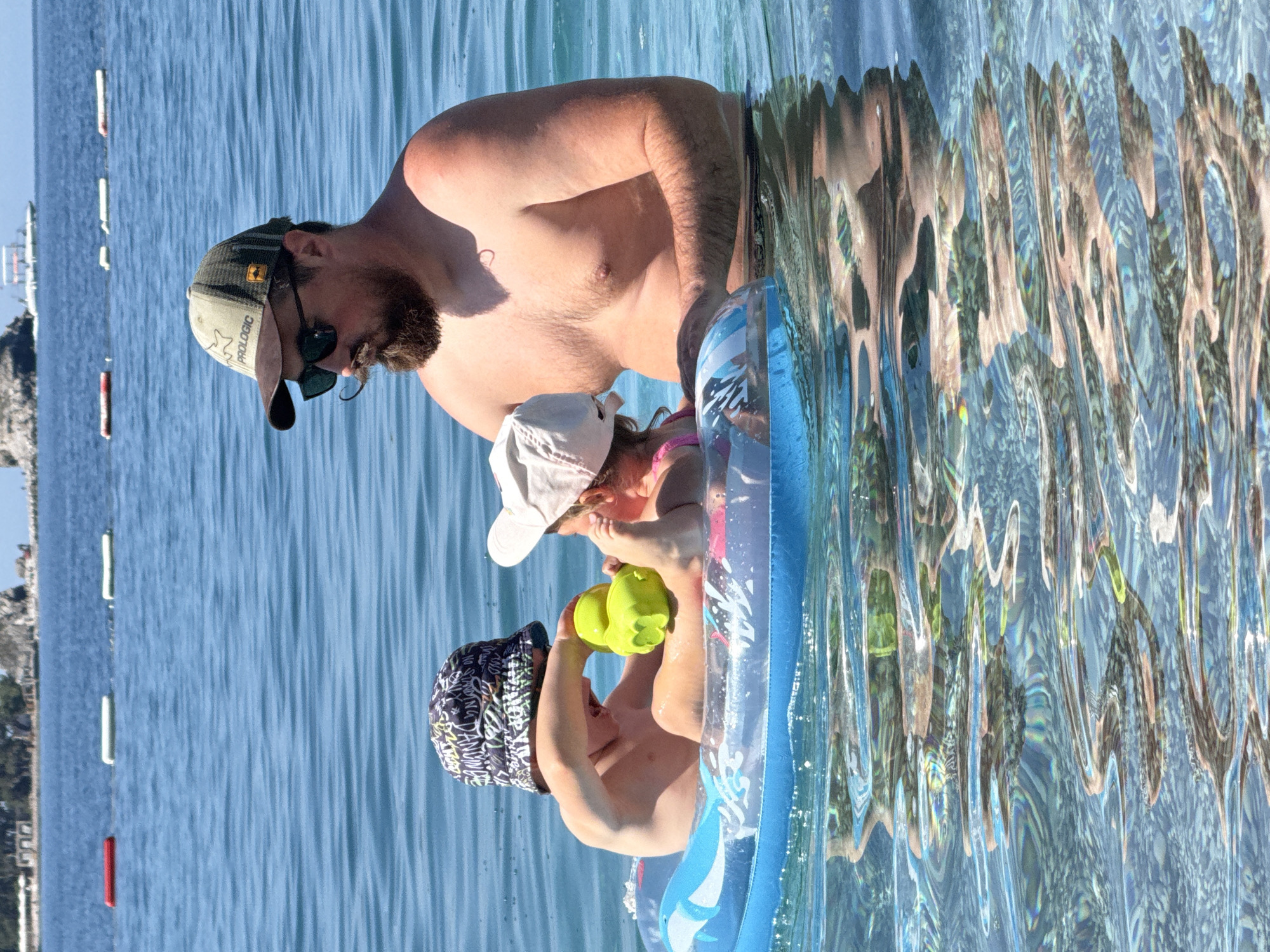 An adult male and two young children are depicted swimming in the clear blue waters of Boreti, Montenegro. The male, an adult with a prominent beard and curled mustache, is wearing a "PROLOGIC" brand baseball cap, dark sunglasses, and is shirtless, standing chest-deep in the water. His left arm is extended towards a blue inflatable ring. Inside the inflatable ring are two young children. The child on the left, a boy, wears a patterned bucket hat and holds a bright yellow, possibly duck-shaped, toy with both hands. The child on the right, younger and wearing a light-colored baseball cap and pink swimwear, is also in the ring, facing away from the camera. The man appears to be supervising and supporting the children. The water is highly transparent, revealing reflections and a pebbly seabed below. In the background, a distant coastline with some landmass and sparse vegetation is visible under a bright sky, along with several red and white cylindrical buoys marking a swimming area and a small white boat further out.