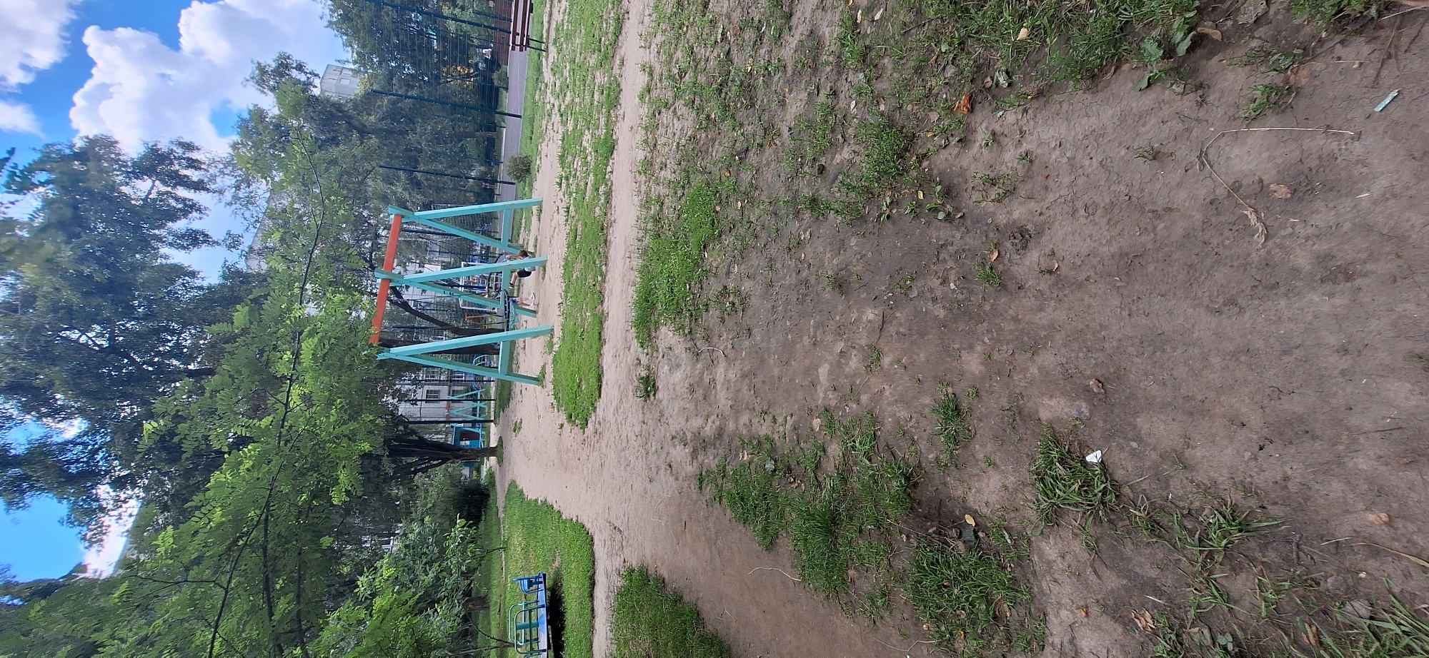 The image shows a park scene in Bila Tserkva, Ukraine. The main subjects are playground equipment, specifically a blue A-frame swing set with orange accents, and a small blue merry-go-round. A person is visible on the swing set, seemingly interacting with it. The setting is an outdoor park with a dirt path winding through it, bordered by patches of grass and trees. In the background, there are buildings and a chain-link fence, suggesting an urban park.
The time of day appears to be daytime, with clear skies and some fluffy white clouds visible. The weather seems pleasant. The overall mood is casual and recreational, typical of a park environment. There are no visible text elements in the image. The image is oriented in landscape mode, with the playground equipment located on the left side and the dirt path dominating the right side. The greenery from the trees and grass adds a natural element to the scene.