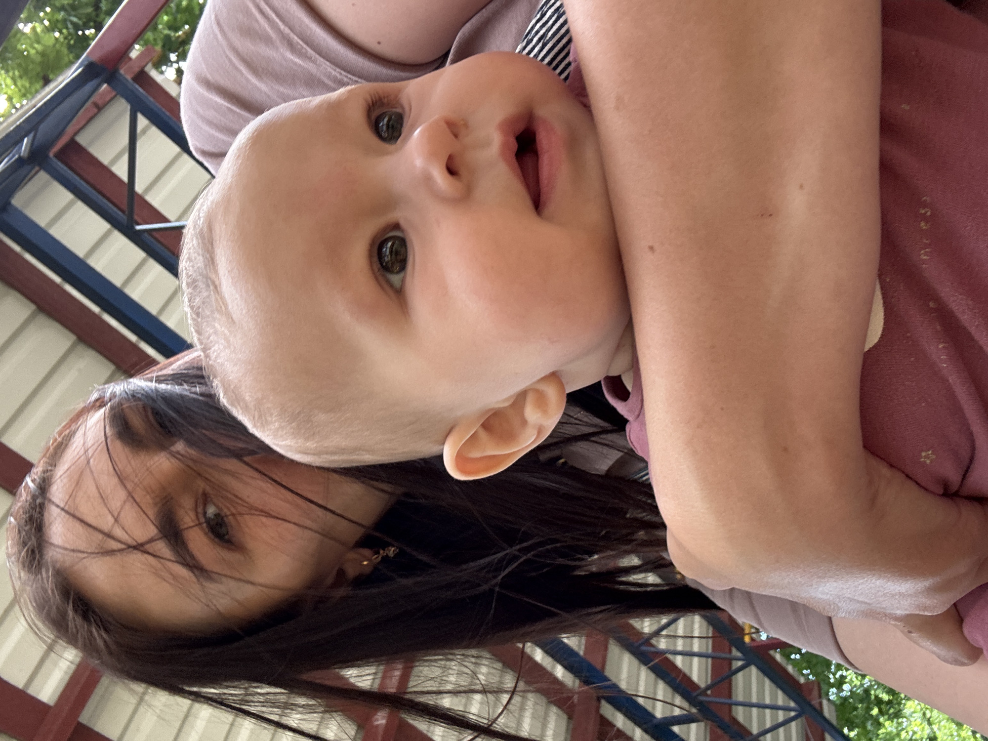 A fair-skinned baby is being held by an individual with dark hair. The baby has light brown eyes and an open mouth. The individual holding the baby is partially visible and has brown hair and dark eyes. The background includes a structure with blue and red beams, along with greenery in the upper right corner. It is impossible to determine the precise location in Sochi, Russia based solely on the image provided.