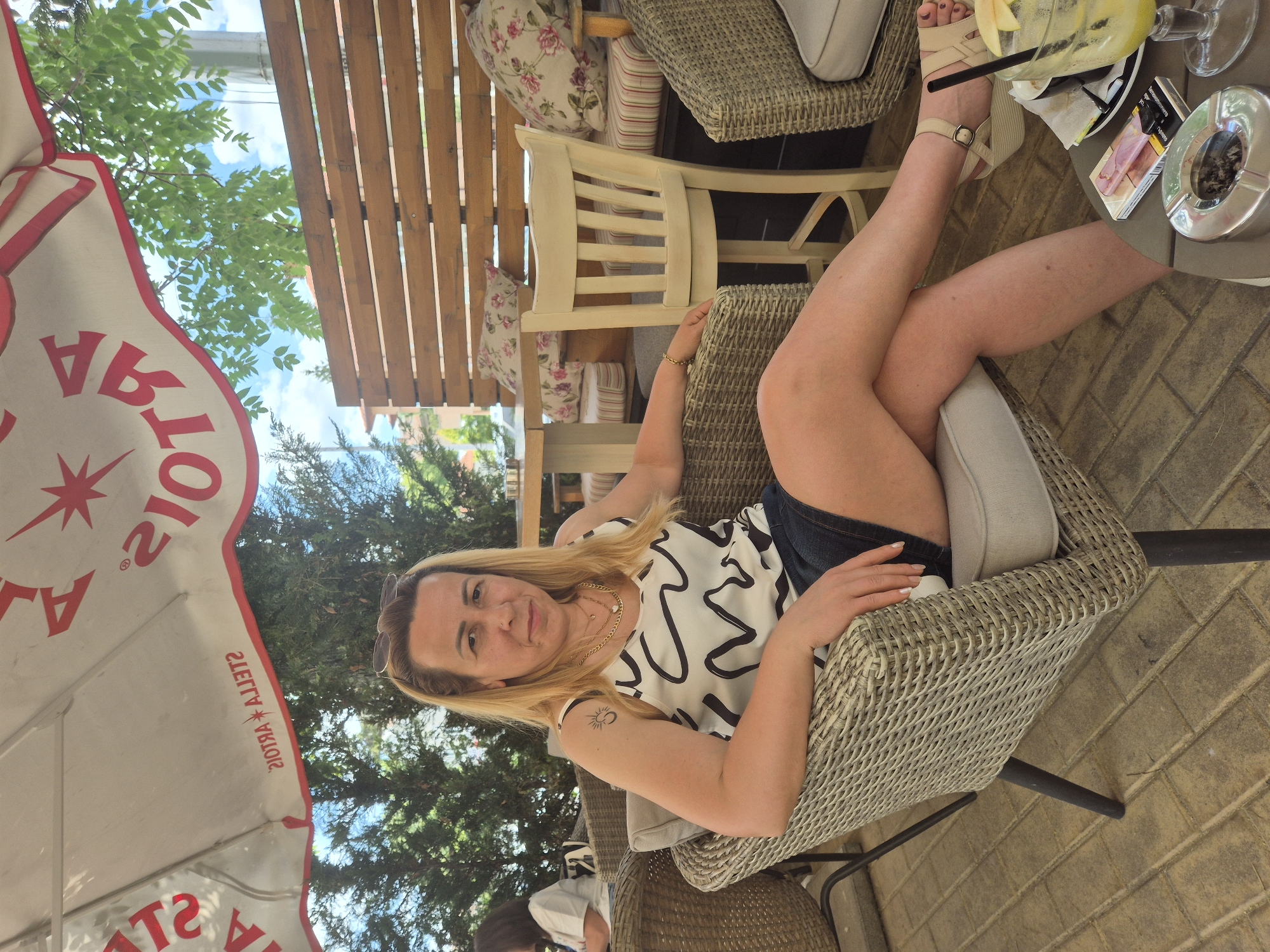 A woman with blonde hair and a white and black tank top is sitting in a wicker chair outdoors. She is wearing denim shorts and tan platform sandals. The woman is looking at the camera with a neutral expression. In front of her is a table with an ashtray, cigarettes, and a glass of yellow liquid with a straw. The background shows a wooden fence, pillows with floral patterns, and trees. The scene appears to be at an outdoor cafe or restaurant.