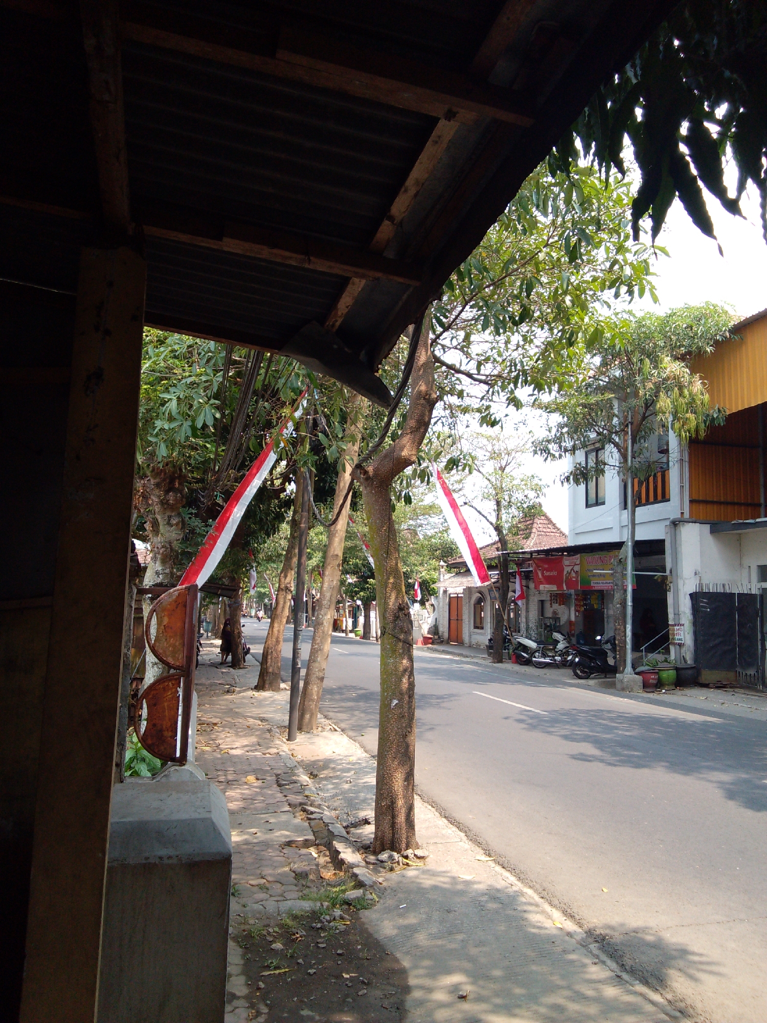 This image depicts a street scene in Trenggalek, Indonesia, possibly during a national holiday or event, as indicated by the presence of Indonesian flags. On the left side of the frame, a weathered, covered walkway is visible, supported by wooden beams and a corrugated metal roof. The walkway has a concrete floor and is lined with trees. Two red and white Indonesian flags are prominently displayed, tied to poles or trees along the sidewalk. Further down the street, buildings can be seen, including a white two-story structure with balconies and a yellow-tiled roof on the right. Several motorcycles are parked on the sidewalk in front of one of the buildings. The street itself is paved and appears to be relatively busy, with trees casting dappled shadows on the asphalt. The environment suggests a warm, sunny day, characteristic of the tropical climate in Indonesia.