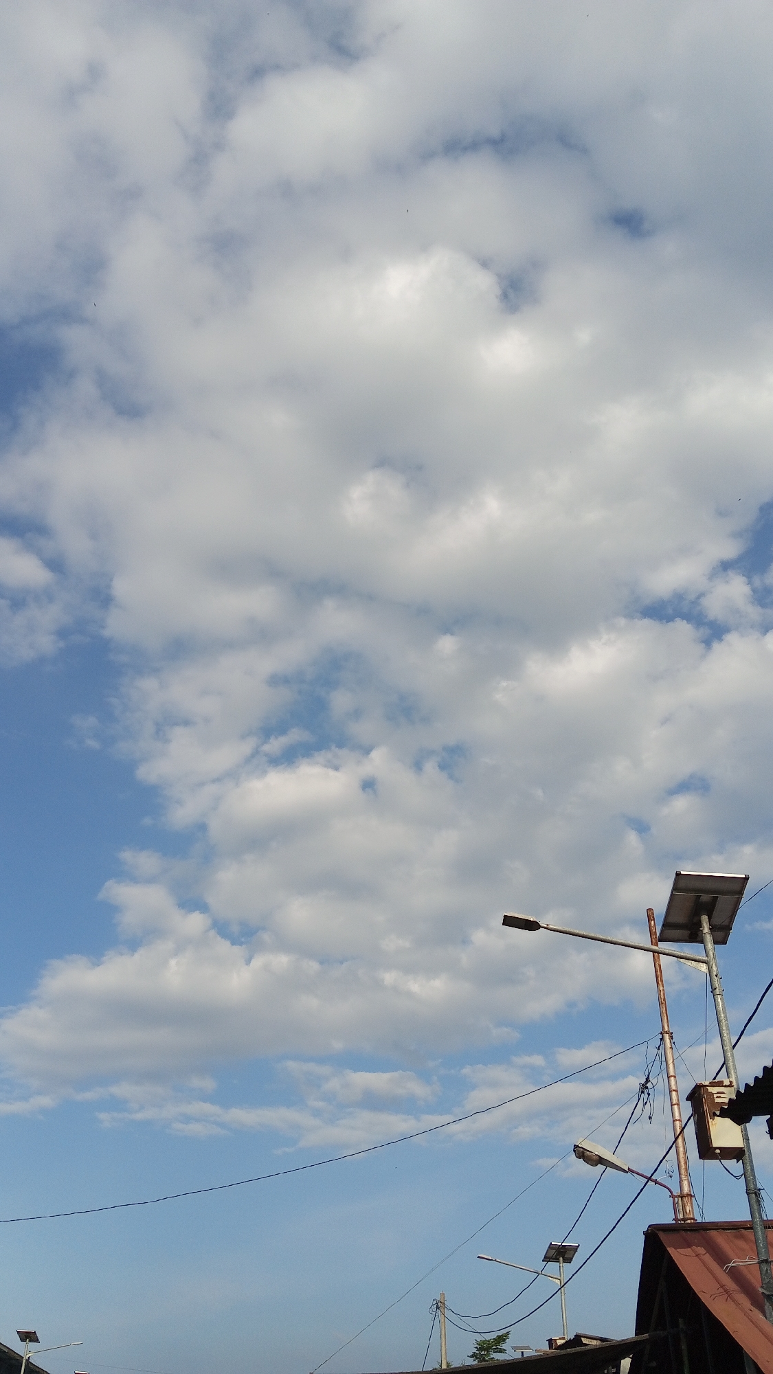 The image shows a cloudy blue sky with a few distant birds. In the foreground, there are several streetlights, some with solar panels attached. Wires are strung between poles, and part of a reddish-brown corrugated roof is visible. The scene appears to be from an outdoor setting, possibly in a residential or urban area, given the presence of streetlights and wires.