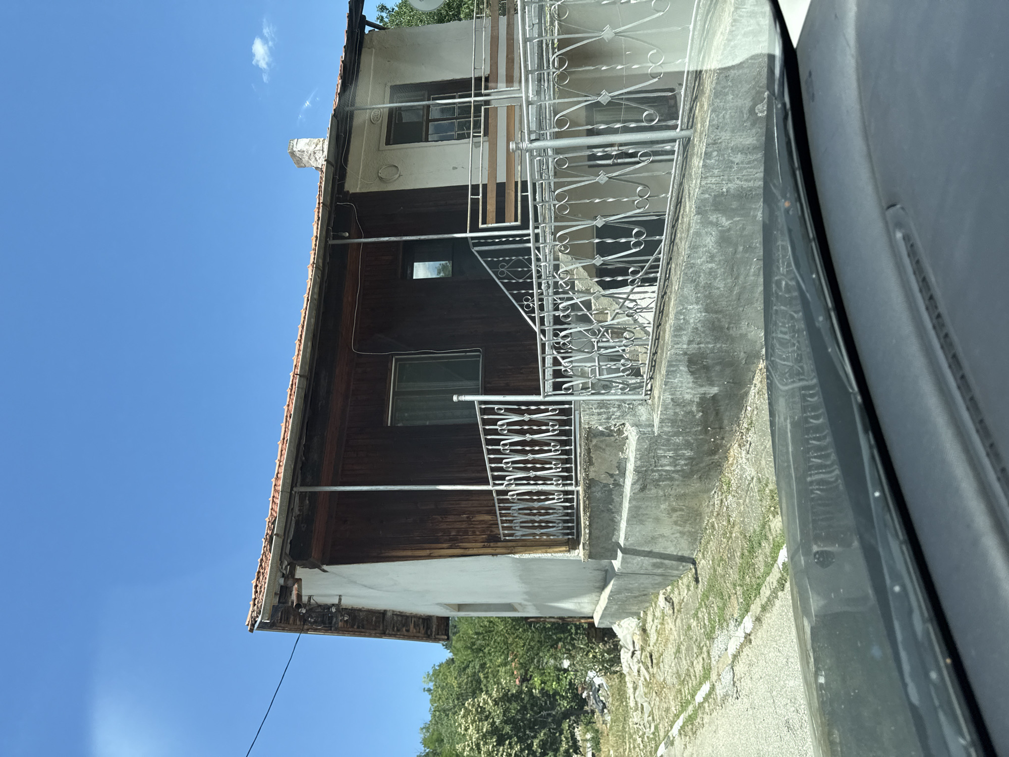The image shows a two-story house on a bright, clear day in Gornoslav, Bulgaria. The house has a white stucco exterior on the right side and a wooden facade on the left. A red tile roof is visible, and a brick chimney stands on the right side of the roof. There are windows on both floors, and a covered porch with a metal railing runs across the front. A set of concrete stairs with a metal railing leads up to the porch. The house is surrounded by greenery, with trees and bushes visible in the background. The foreground shows the dashboard and windshield of a vehicle, suggesting the photo was taken from inside a car. The area appears to be rural, with a dirt or gravel road leading up to the house.