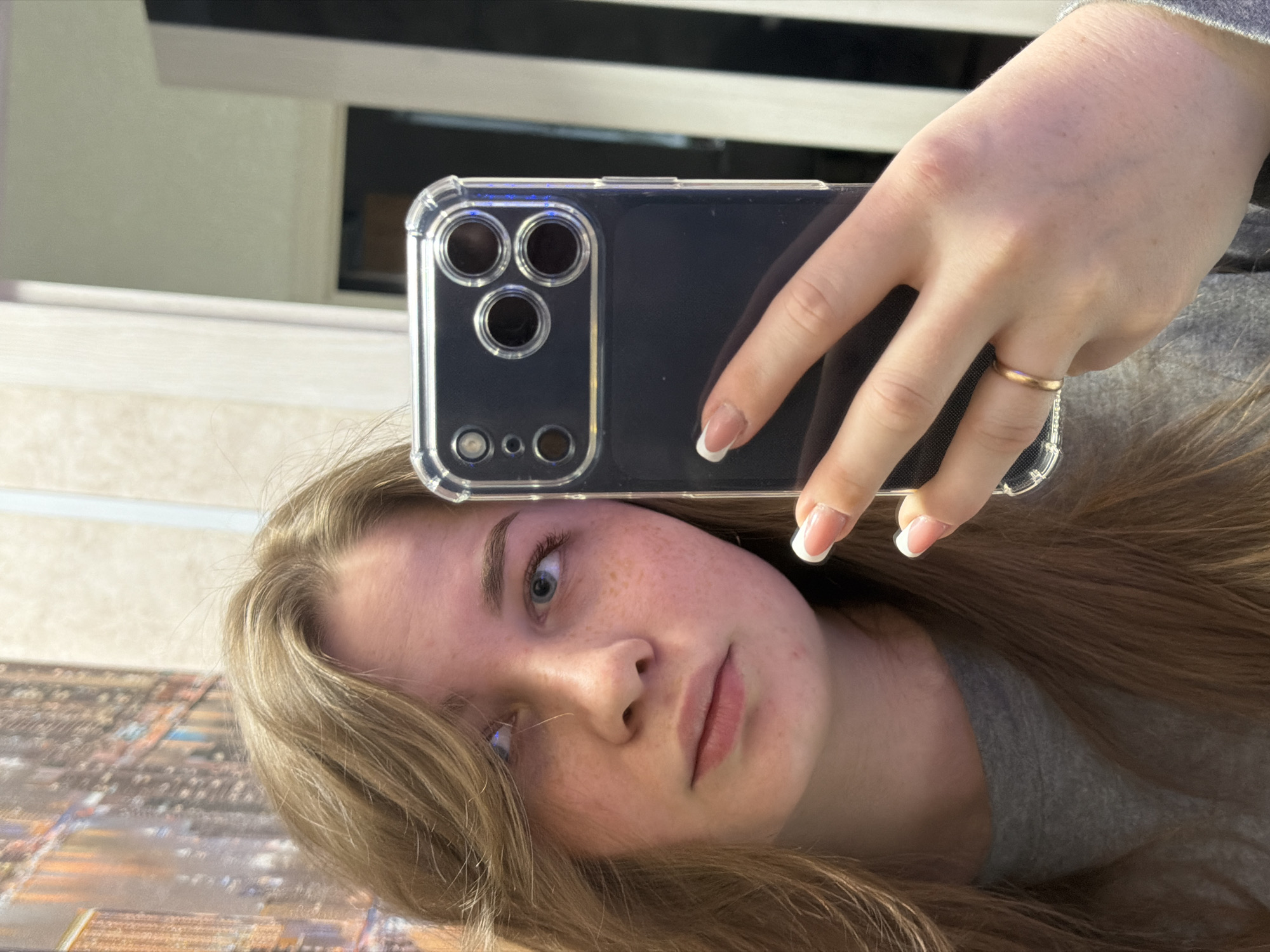 A selfie taken by a fair-skinned young woman with light brown hair and blue eyes. She's holding a black smartphone with a clear protective case. The phone's camera lens configuration is visible. She is wearing a simple gold ring on her ring finger, and has a white-tipped manicure. She is wearing a gray shirt. In the background, out of focus, are a wall, a piece of furniture, and what appears to be an image or decoration suggesting a city scene; the lighting suggests an interior location. There is no indication or suggestion that this photo was taken in Большой Букор, Russia, or any specific location for that matter.