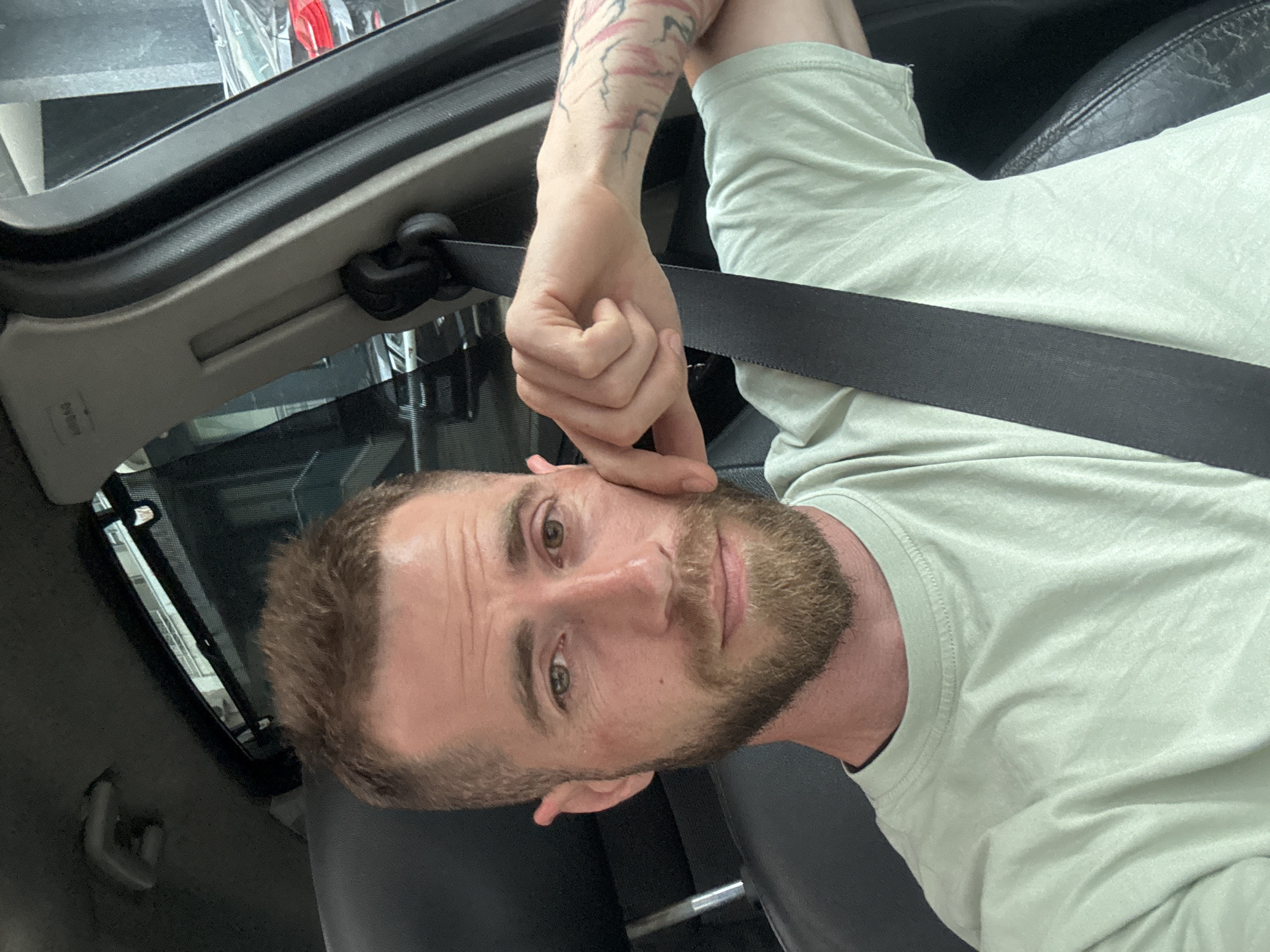 A man is the central subject of this selfie, captured inside a car in Budva, Montenegro. He has a medium-length beard and short brown hair, with light-colored eyes. He wears a light green long-sleeved t-shirt. Tattoos featuring abstract, branch-like patterns in red and pink hues are visible on his left forearm. His left hand is raised, with fingers touching his left cheek, giving him a thoughtful or relaxed expression as he looks directly into the camera.
He is secured by a black seatbelt across his chest. The car's interior is visible, featuring a beige or light grey A-pillar on the left, where the word "AIRBAG" is distinctly marked. Black car seats and a dark headliner are also part of the setting. Bright daylight illuminates the interior, suggesting the photo was taken during the day. The overall mood is casual and direct.