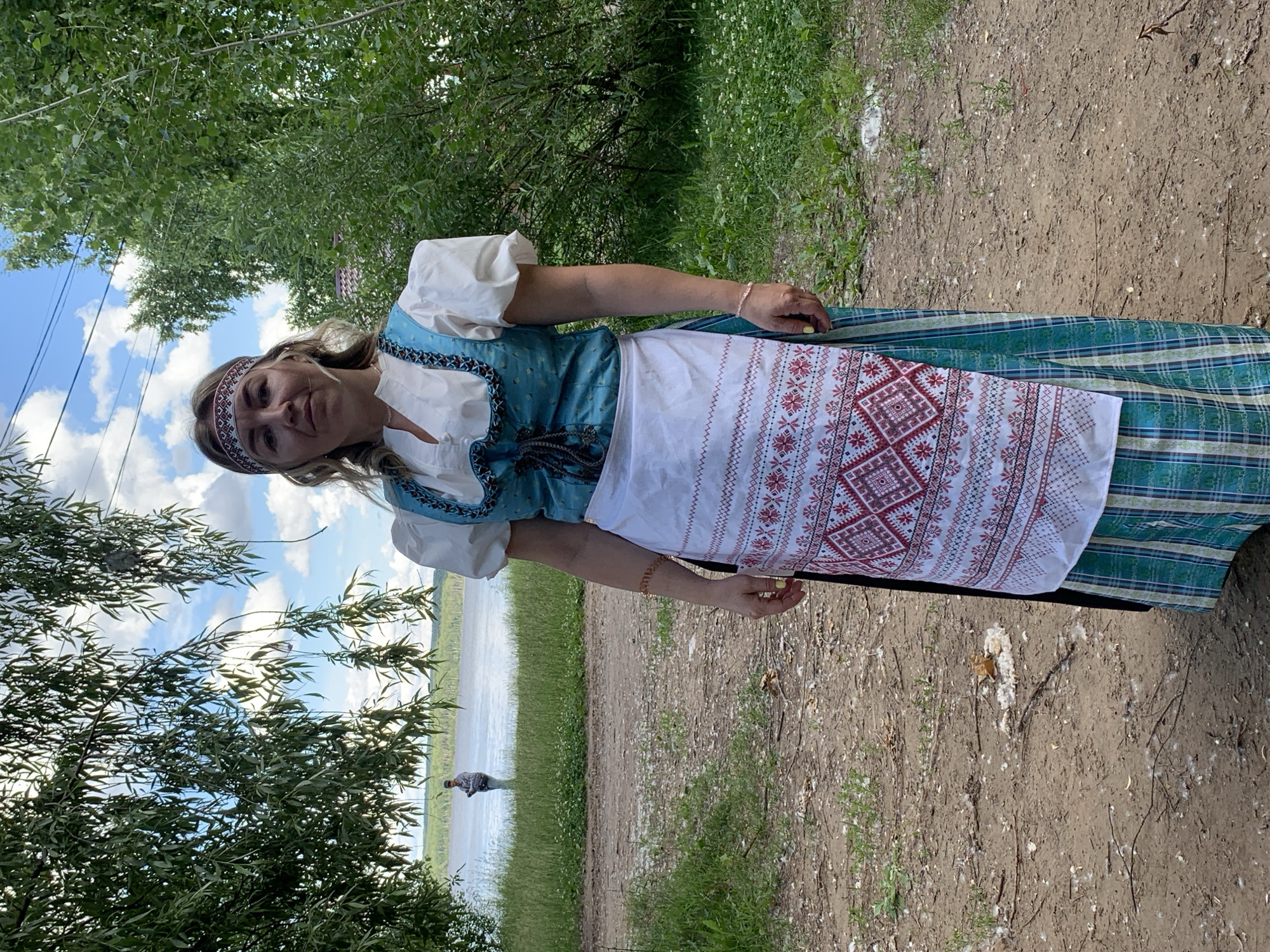 A woman is pictured wearing traditional Russian attire, including a white blouse with puffed sleeves, a blue embroidered vest, and a white apron with red and white embroidery. She also wears a patterned headband and a plaid skirt. She is standing outdoors in what appears to be a park or natural area. Behind her, a body of water can be seen, possibly a lake or river, with a treeline in the distance. The sky is blue with scattered clouds. To the left of the woman, leafy green trees are visible. The ground in the foreground is a dirt path with some sparse vegetation. A person is visible in the distance walking near the water. The lighting suggests it is daytime, likely midday, with bright sunlight. The overall scene evokes a sense of a cultural or festive event, or simply a person enjoying the outdoors in traditional dress.