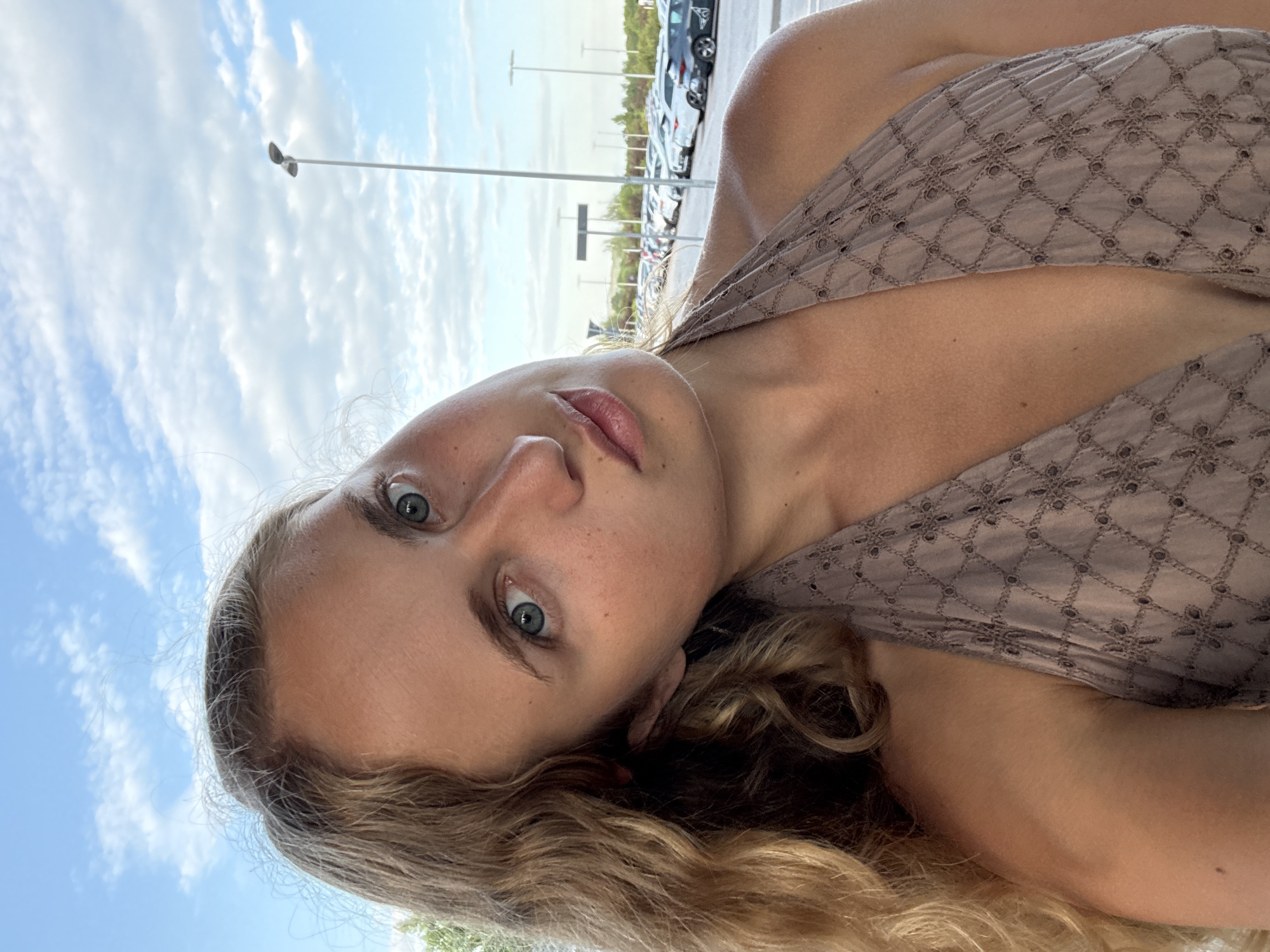 The image shows a young woman with blonde, wavy hair, taking a selfie outdoors. She is wearing a light brown halter top with a geometric pattern. Her head is tilted to the side, and she is looking directly at the camera with blue eyes. Her lips are slightly parted.
In the background, there is a parking lot filled with cars, with many vehicles parked in rows under a bright blue sky with scattered white clouds. A tall, thin streetlamp is visible on the right side of the frame, and further in the distance, more streetlamps and some vegetation can be seen. The setting appears to be an outdoor parking area, possibly near an airport or a large commercial establishment, consistent with locations found in Ronchi dei Legionari, Italy. The lighting suggests it is daytime, likely late afternoon or early evening due to the angle of the light.