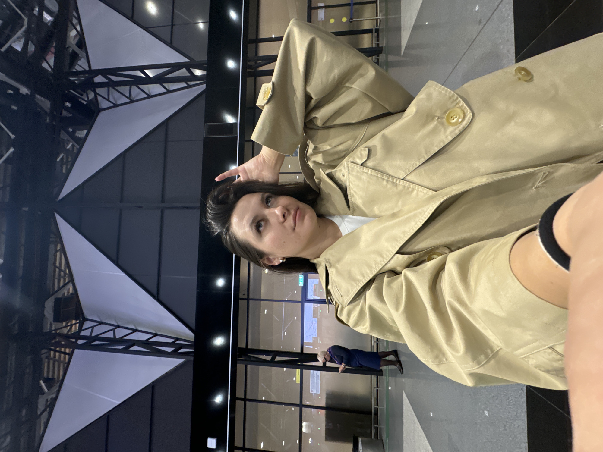 A woman in a tan trench coat poses indoors at Sheremetyevo International Airport in Khimki, Russia. The background features a high ceiling with triangular architectural elements, glass windows, and other people in the distance.