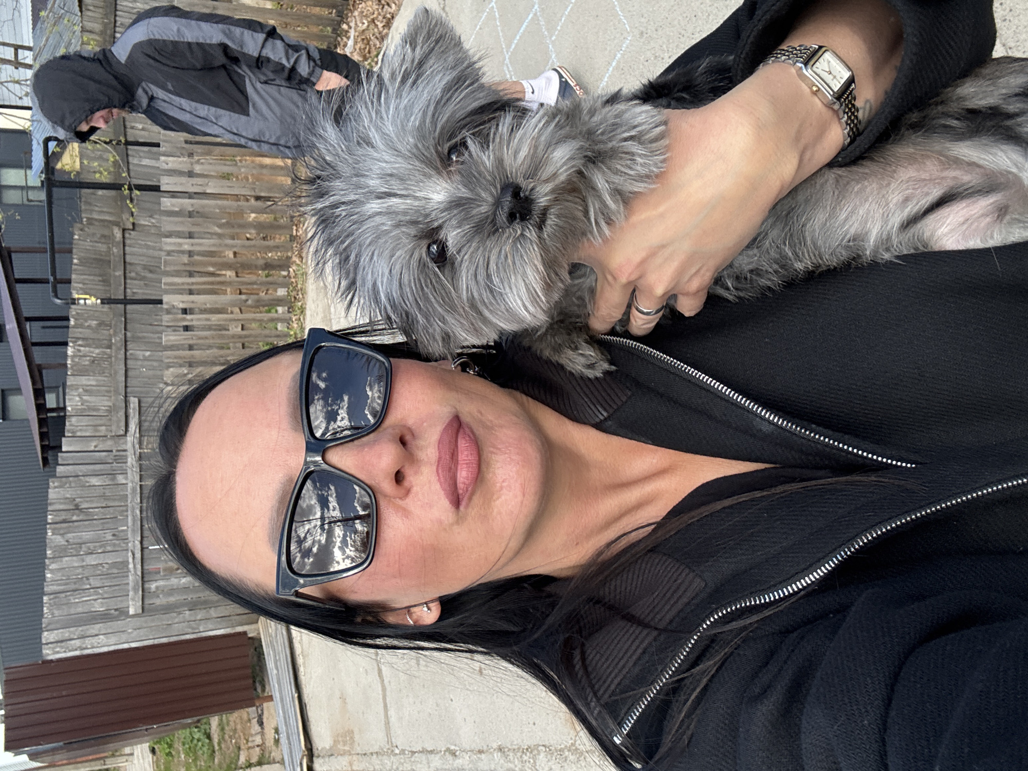 The image is a selfie of a woman in a black jacket and sunglasses, holding a gray, shaggy small dog. The sunglasses reflect the sky with clouds. The woman is also wearing a watch. A man wearing a black and gray jacket with a hood is partially visible in the background. The location is likely outdoors, with a wooden fence and concrete visible. The setting looks residential. It's not possible to definitively confirm the city of Chaikovsky, Russia without additional information or identifying landmarks within the image.