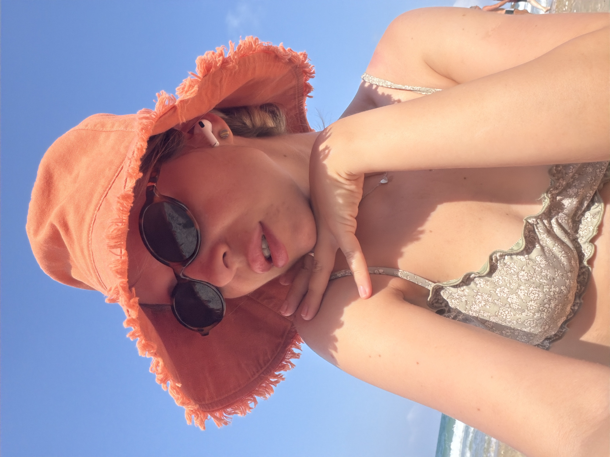 A woman, wearing an orange bucket hat with frayed edges, dark sunglasses, and a patterned bikini top, poses for a selfie on a beach in Bat Yam, Israel. She has light skin, her hands are resting on her chin, and she is wearing white wireless earbuds. The background shows a clear blue sky and a body of water with small waves.