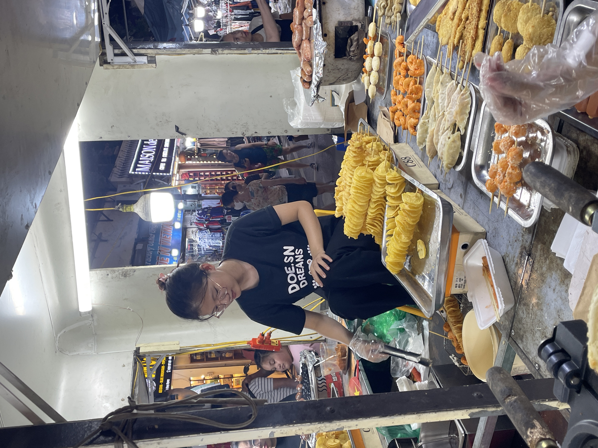 This is a street food stall in Hanoi, Vietnam, likely at a night market or busy commercial street. The stall is filled with various fried snacks. In the foreground, a young woman wearing glasses and a black t-shirt with white text that reads "DOE.SN DREAMS" is preparing food. She is wearing disposable gloves and using tongs to handle a skewer of food. To her left, a tray of spiral-cut fried potatoes is prominently displayed, along with other fried items. Behind her, a young child in a pink shirt looks on. Further back, other people are visible browsing the stalls, with clothing and other merchandise displayed. The environment appears to be an outdoor market with artificial lighting illuminating the scene. The overall impression is a vibrant and bustling food market.