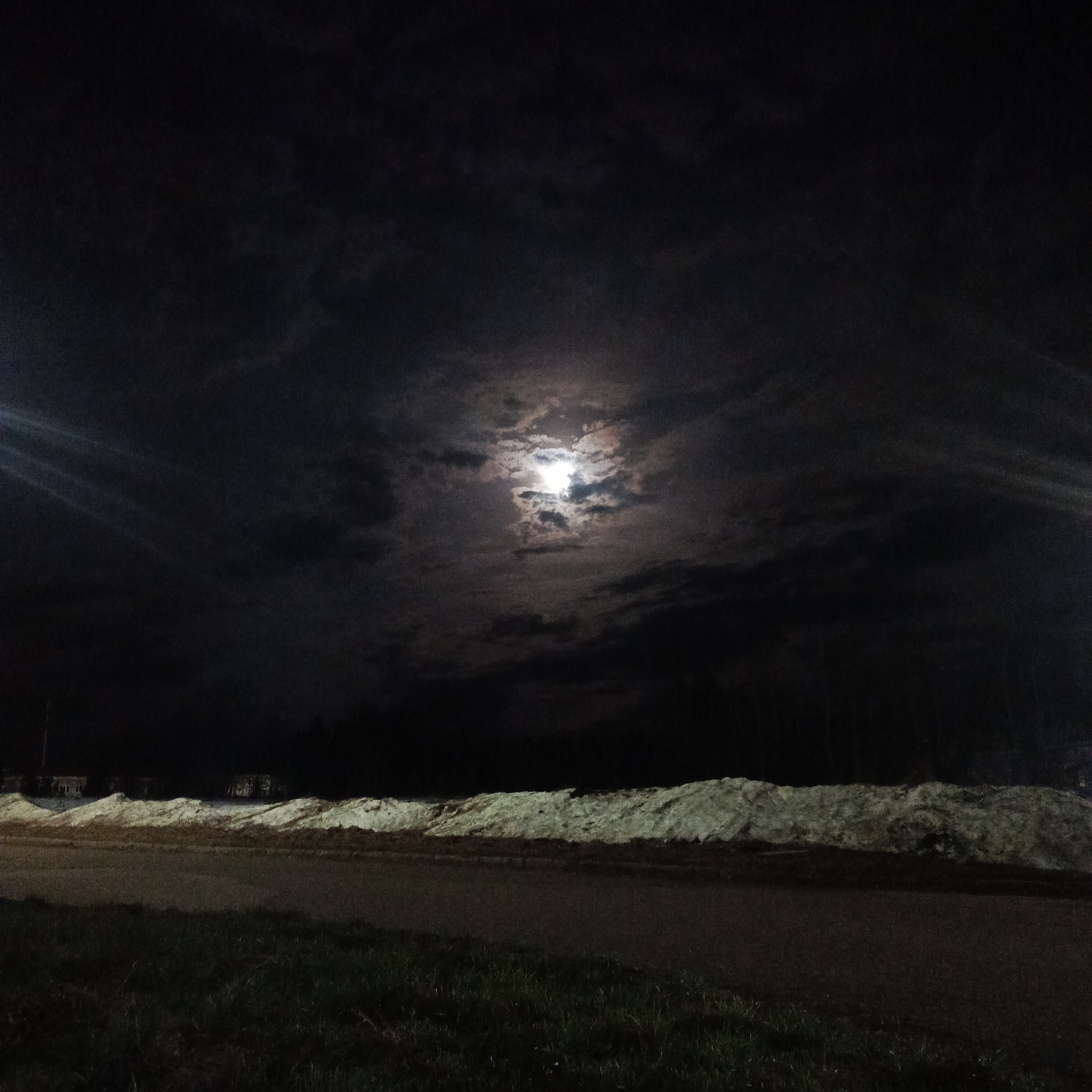A night scene in Марковский, Russia. A road runs horizontally across the lower portion of the frame. A grass-covered median separates the road from a large pile of snow. In the background, trees and buildings are visible. The sky is dark and cloudy with a bright full moon. Lens flares emanate from the moon.