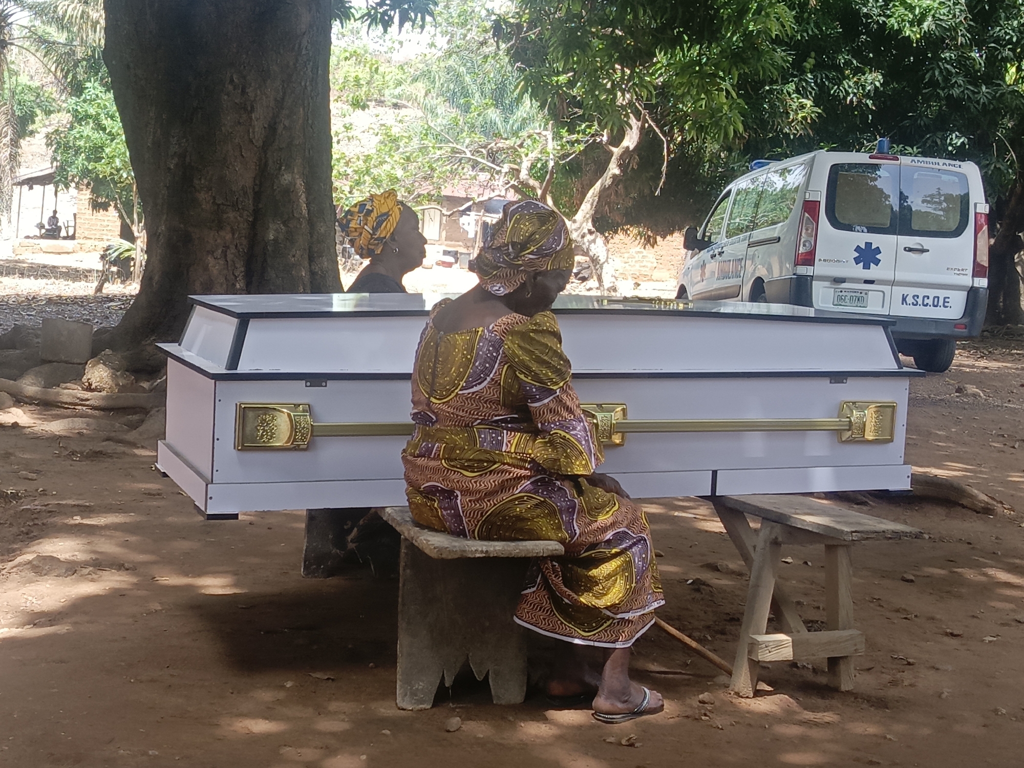The image depicts a solemn scene in Ninzo South, Nigeria. In the foreground, an elderly woman dressed in a patterned brown and gold outfit sits on a low concrete stool. Her back is to the viewer, and she rests her arms on a white casket with gold handles. Beside her is another woman wearing a yellow headwrap whose face is partially visible. Behind the casket is a parked white ambulance with the words "AMBULANCE" and "K.S.C.O.E." visible. The scene is shaded by large trees. The ground is covered in reddish-brown earth. In the distance, a small structure is visible.