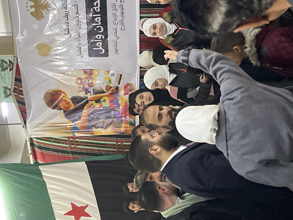 An indoor scene in Kafr Batna, Syria, depicts a dense gathering of approximately 20-30 individuals, primarily adults. In the immediate foreground, an individual wearing a grey coat and a white head covering is seen from behind, with their right arm raised vertically above the crowd. To their right, the back of a child's head, wearing a brown hood with fur trim, is visible.
The mid-ground features a tightly packed group of men and women. Several men with beards are present, one in a dark suit jacket, while numerous women wear hijabs; some are observing attentively, others are smiling or engaging with unseen activity.
The background displays a large flag on the left, characterized by green, white, and black horizontal stripes with a red five-pointed star centered on the white stripe (the Syrian opposition flag). Adjacent to this, a large white banner features a graphic of a smiling child playing with colorful building blocks and toys. Arabic text on the banner identifies it as relating to the "Damascus Countryside Governorate," a "Women and Child Affairs Department," and phrases translating to "Safety and Hope." Two golden eagle emblems are visible on the upper right of this banner. Behind the banner, striped fabric in red, black, and white is discernible, suggesting decorative drapery or a tent structure. Overhead lighting fixtures are partially visible at the top of the frame.