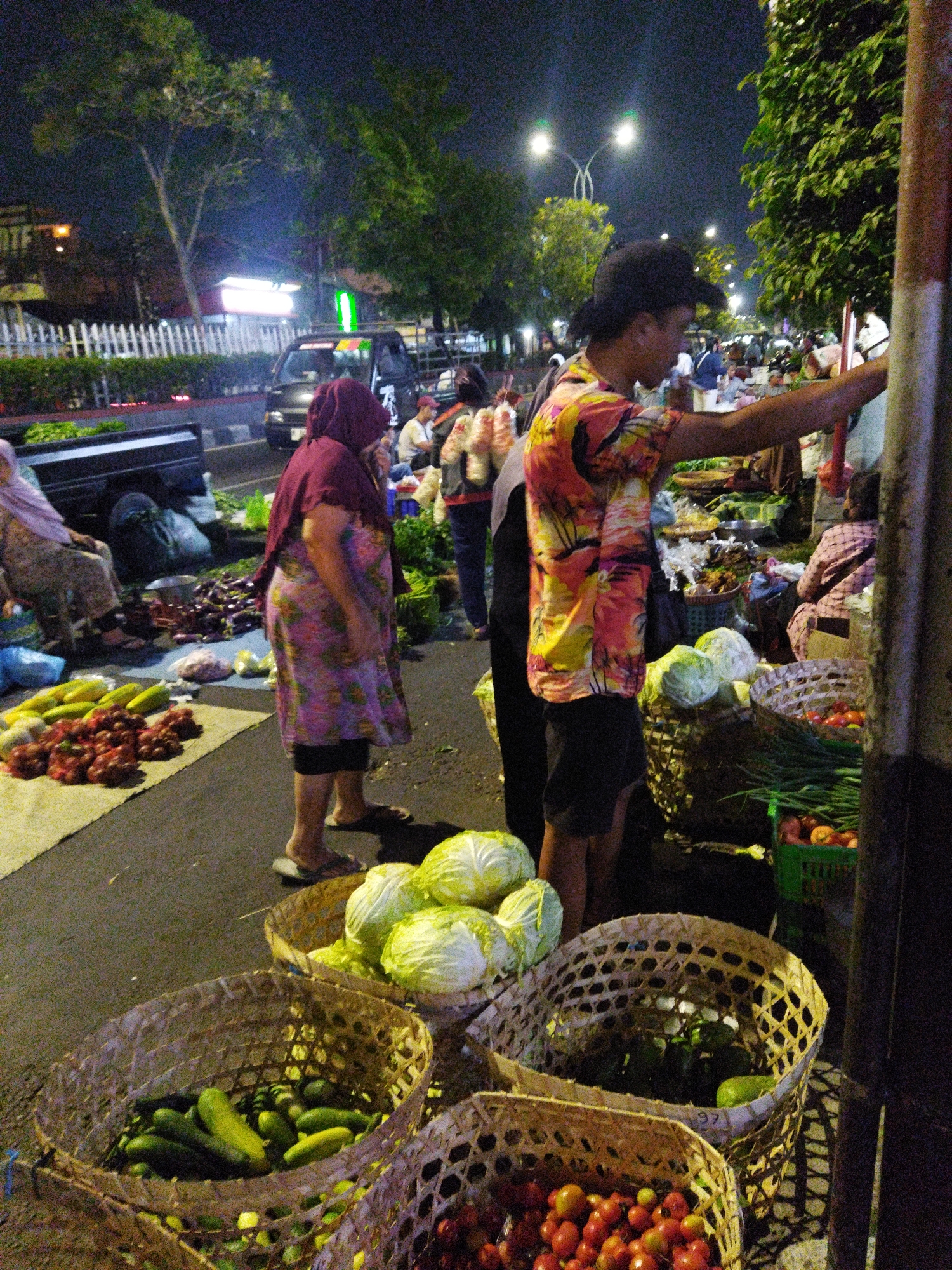A street-level view captures a nighttime scene at a market, likely in Semarang, Indonesia, given the prevalence of tropical produce and street vendor setups. The foreground features several woven baskets brimming with fresh vegetables, including cucumbers and what appear to be tomatoes. A woman in a purple headscarf and a patterned dress stands on the right, facing away from the camera, and appears to be a vendor or customer. To her left, a man in a black hat and a brightly colored, tropical-patterned shirt is engaged in an activity at a market stall, possibly a vendor arranging produce. Behind them, more market stalls are visible, with various vegetables, including cabbages and eggplants, displayed in baskets and on the ground. A black vehicle and streetlights are visible in the background, suggesting an urban setting. The overall atmosphere is that of a bustling local market operating after dark.