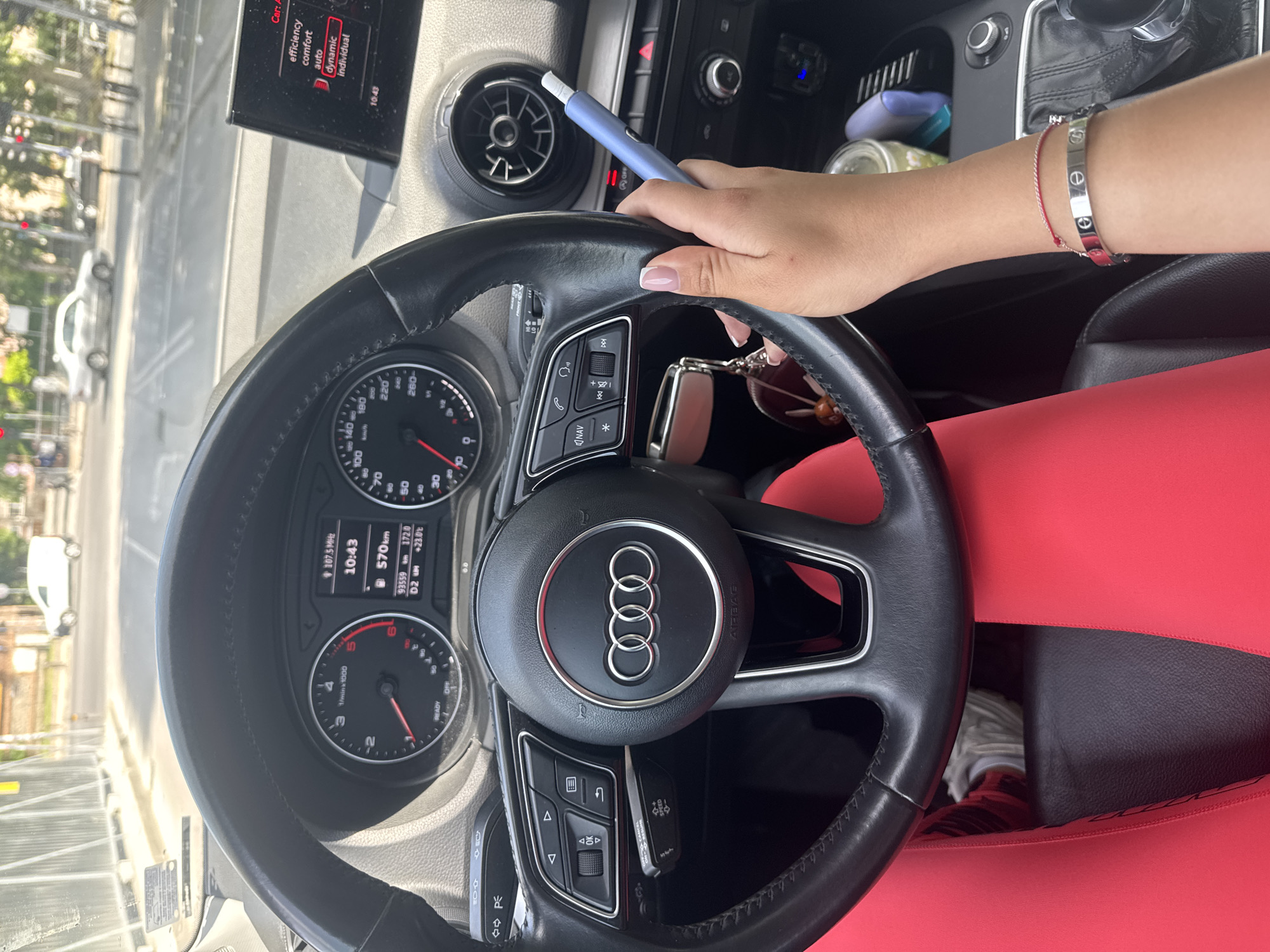 The image shows the interior of an Audi car, viewed from the driver's seat. A person's hands are visible; one hand rests on the steering wheel, while the other holds a blue electronic cigarette. The dashboard displays the time as 10:43, the radio station as 107.5 MHz, and the mileage as 93559 km. The car's infotainment screen shows driving mode options: "efficiency," "comfort," "auto," "dynamic," and "individual," with "dynamic" currently selected. Outside the car, a street scene is visible through the windshield, with cars and buildings suggesting an urban environment. The person is wearing bright red leggings and a silver bracelet.