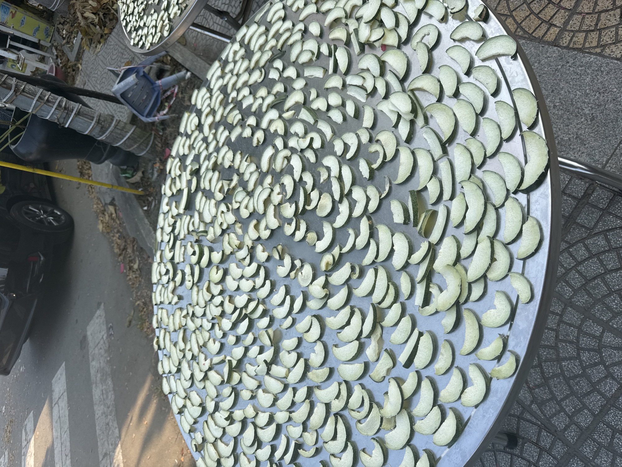 The image shows a close-up, high-angle view of a large, round, silver metal table densely covered with numerous slices of a light green fruit, likely some variety of melon or squash, laid out for drying. The slices are crescent-shaped, with a visible rind and pale interior. The table is situated outdoors on a paved surface, possibly a sidewalk, next to a street.
In the background to the left, part of a car and a bicycle are visible, along with various pieces of discarded debris. There is another, similar table partially visible in the upper-left corner, also covered with slices of the same fruit.
The location is suggested to be Đà Nẵng, Vietnam.