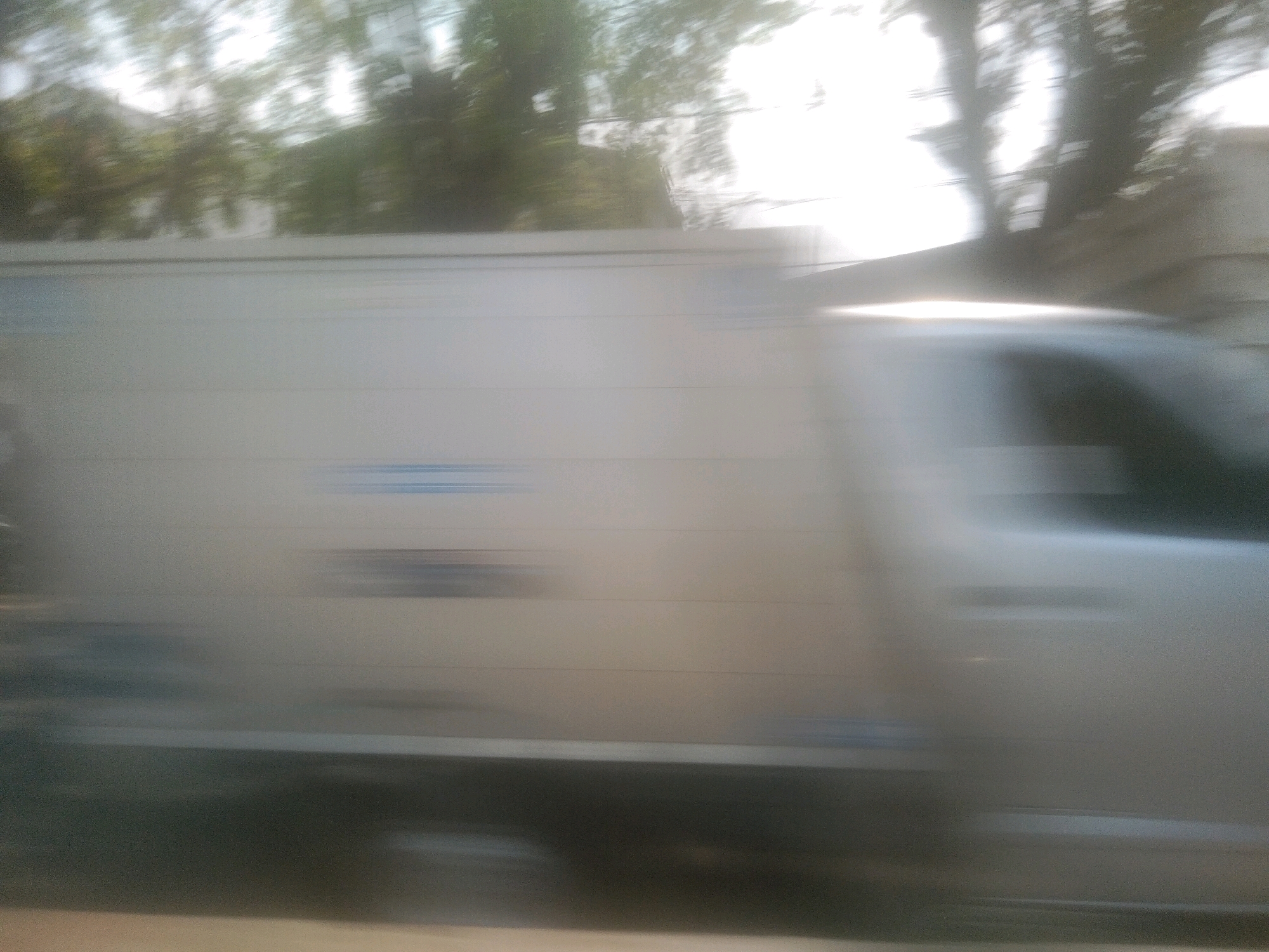 The image captures a blurred view of the side of a white box truck in motion, likely taken from a passing vehicle on a street in Bandar Lampung, Indonesia. The truck's surface is a light, metallic color, with horizontal panel lines visible. Faint blue markings or graphics are discernible on the side of the truck. The background shows a motion blur of trees and buildings, suggesting an urban or suburban environment. The lighting indicates it is daytime, with bright sunlight creating lens flare and washes of light on the scene. The overall impression is one of speed and transit, with minimal sharp details due to the motion blur. No individuals or specific landmarks are clearly identifiable.
