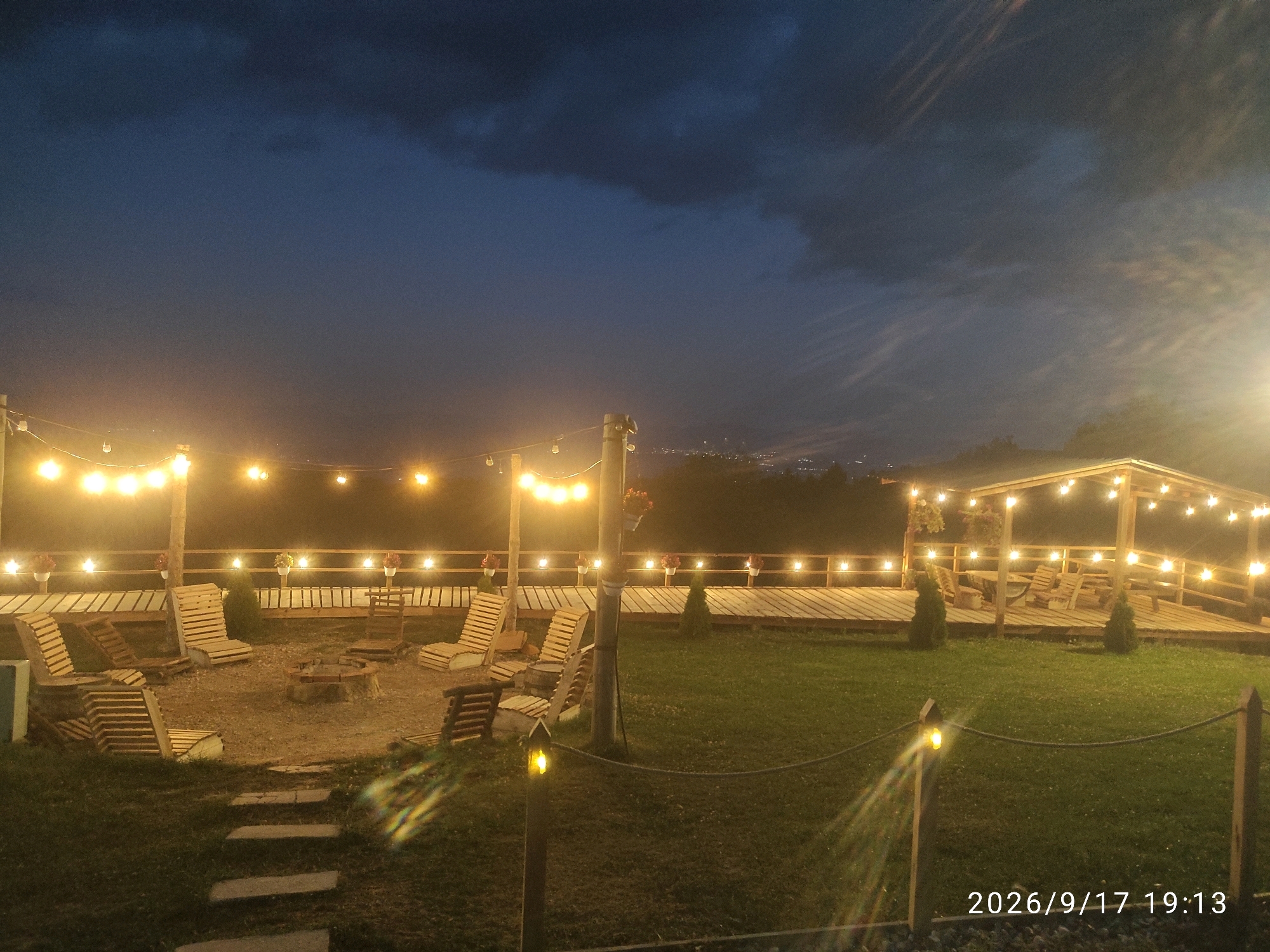 The image depicts an outdoor seating area at dusk, likely in Krushevo OT, North Macedonia. The scene features a fire pit surrounded by wooden lounge chairs, a wooden deck, and a covered patio area. String lights illuminate the area, casting a warm glow on the surroundings. The sky is dark blue with some clouds, indicating the transition from day to night. The overall atmosphere is relaxed and inviting, suitable for outdoor gatherings.