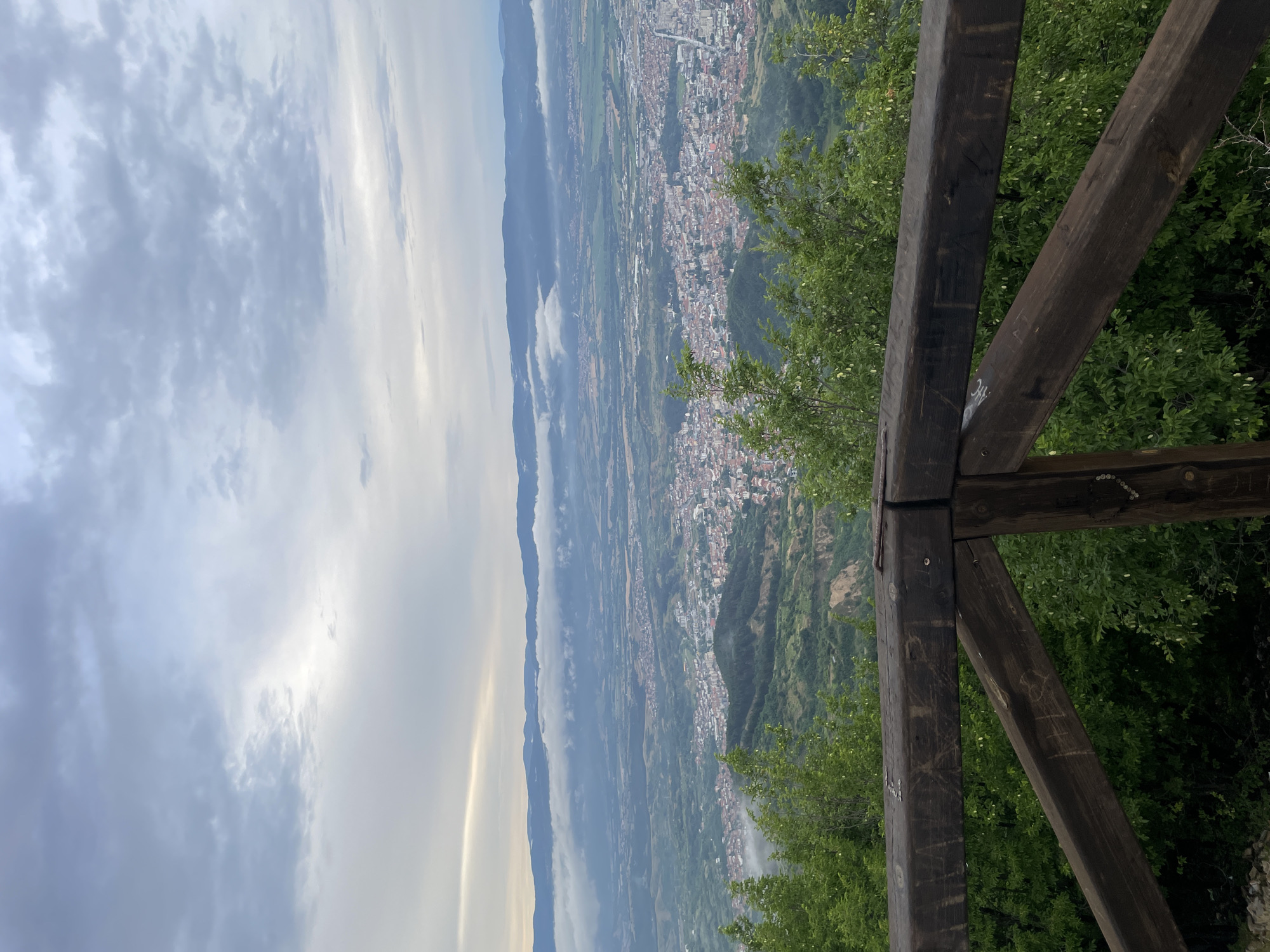 An elevated, wide-angle shot captures a scenic overview of a city nestled in a valley, viewed from a wooden railing. The sky above is overcast with diffused light, indicating either early morning or late afternoon. In the foreground, a weathered wooden railing made of thick, dark timbers forms a protective barrier. Some of the timbers bear graffiti and carvings. Lush green trees with light-colored blossoms are visible behind and around the railing.
The mid-ground reveals a sprawling urban landscape with densely packed buildings featuring red tile roofs. The city extends into the valley, interspersed with patches of green. Rolling hills and mountains form the backdrop, with a layer of low-lying clouds or fog partially obscuring the distant mountain range. The overall impression is one of a peaceful, vast panorama overlooking a settled area. The specific location is not identifiable from the visual information alone; it is a generic representation of a city in a valley surrounded by nature.