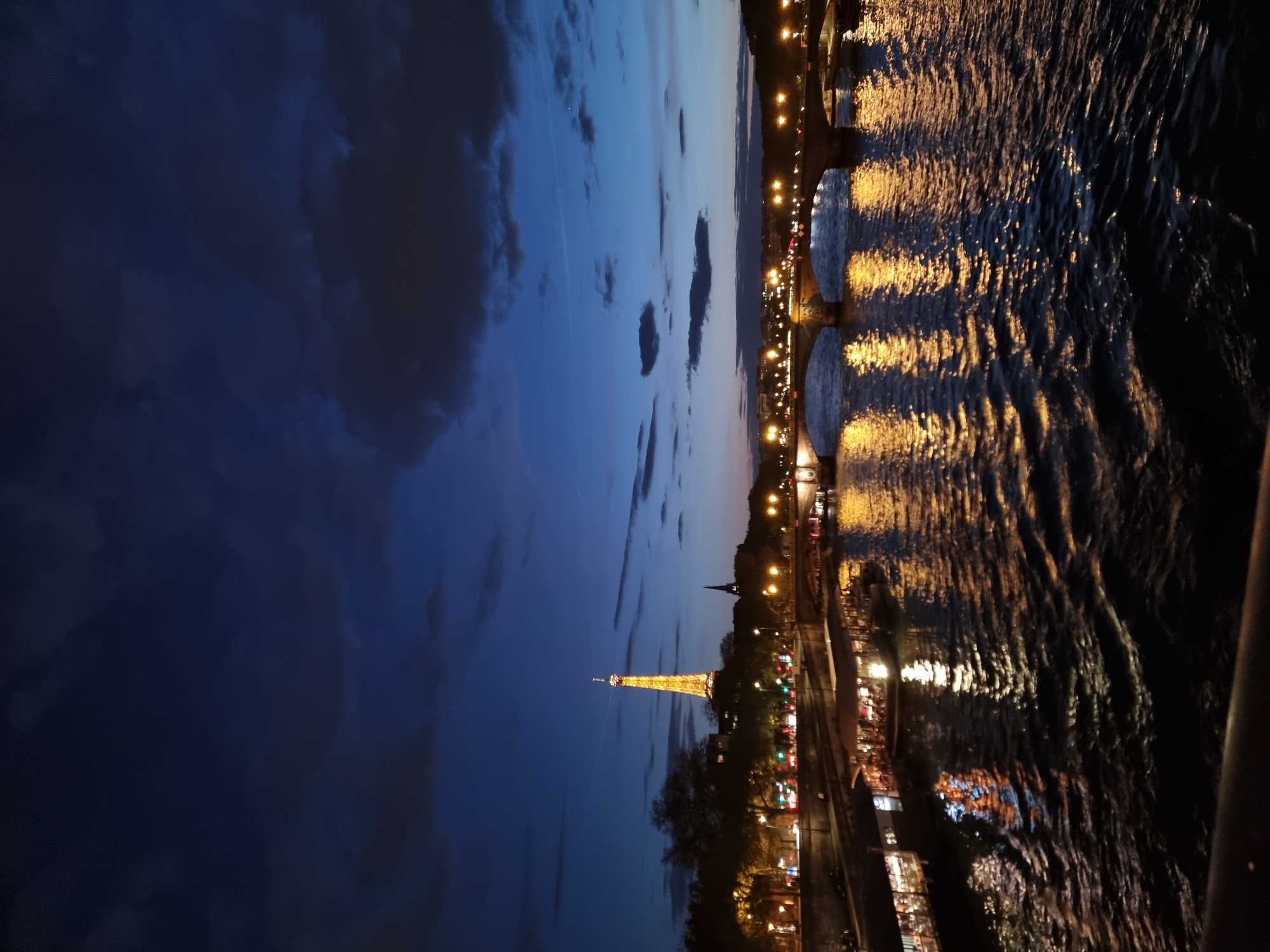 The image is a nighttime view of the Eiffel Tower in Paris, France. The tower is illuminated and stands prominently against a dark blue sky with scattered clouds. Below the tower, city lights create a bright glow along the Seine River. The river reflects the city lights, creating a shimmering effect. In the foreground, the Pont Neuf bridge spans the river, its arches visible and lit. The image has a slight tilt.