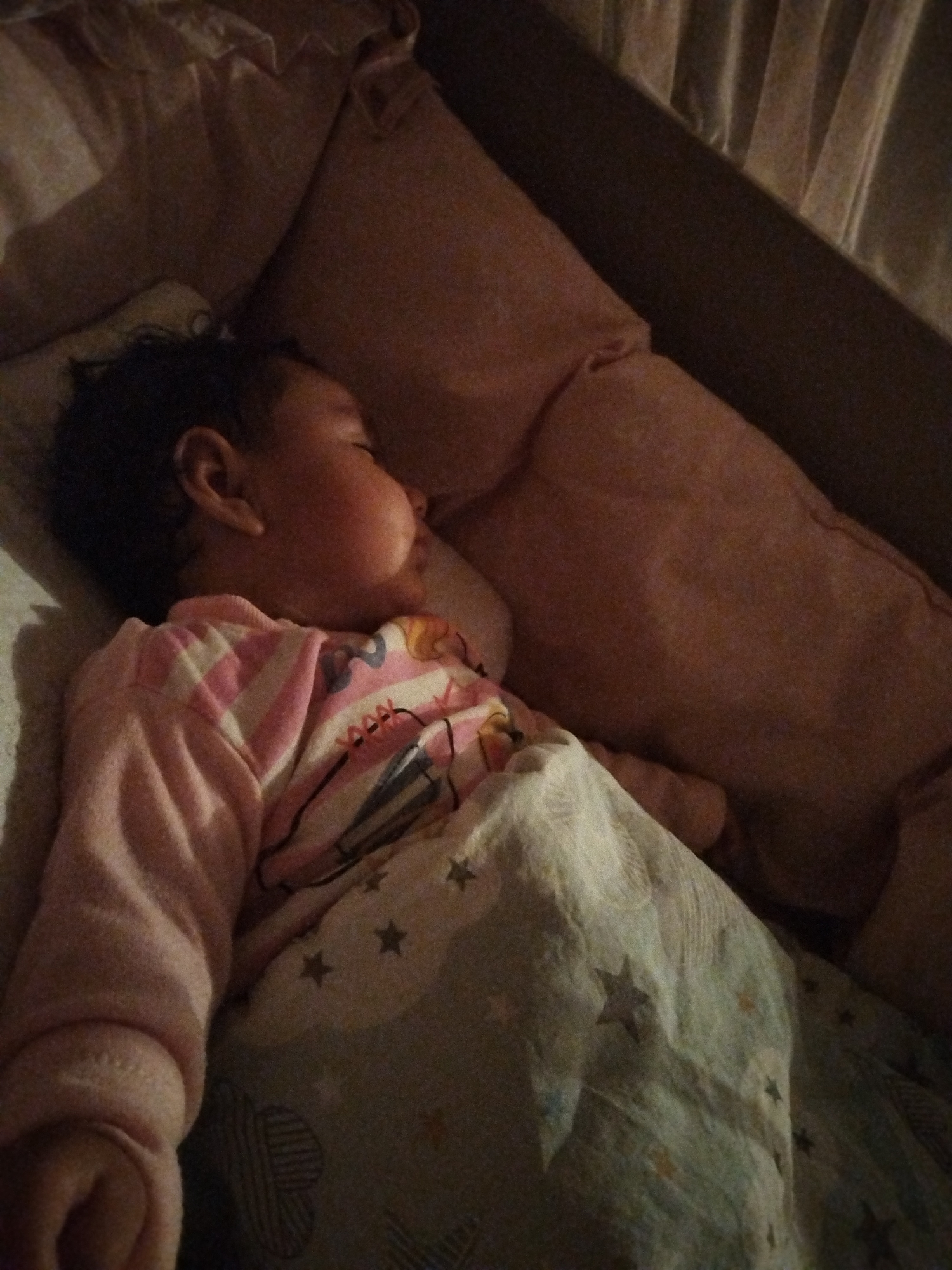 The image captures a close-up, dimly lit view of a baby or toddler sleeping peacefully. The child is positioned on their side, facing slightly away from the camera, with their head resting on a light peach-colored pillow. Their eyes are closed, and their facial features appear soft and relaxed, indicating a deep sleep.
The child is wearing long-sleeved pajamas or a shirt. The sleeves are pink with white horizontal stripes, while the body of the shirt features a graphic design with various colors, including pink, blue, and black outlines, possibly depicting cartoon characters or shapes.
A light-colored blanket, possibly white or pale blue, covers the child from the chest down. This blanket is adorned with a pattern of dark and light stars, along with some circular and striped motifs. Another part of the bedding, possibly a duvet or another pillow, is visible behind the child's head, appearing in a similar peach or light pink hue.
The setting appears to be a bedroom or nursery, likely at night or in a very dimly lit room, creating a serene and quiet atmosphere. The lighting is soft and warm, casting gentle shadows and enhancing the sense of tranquility. No text is visible in the image.