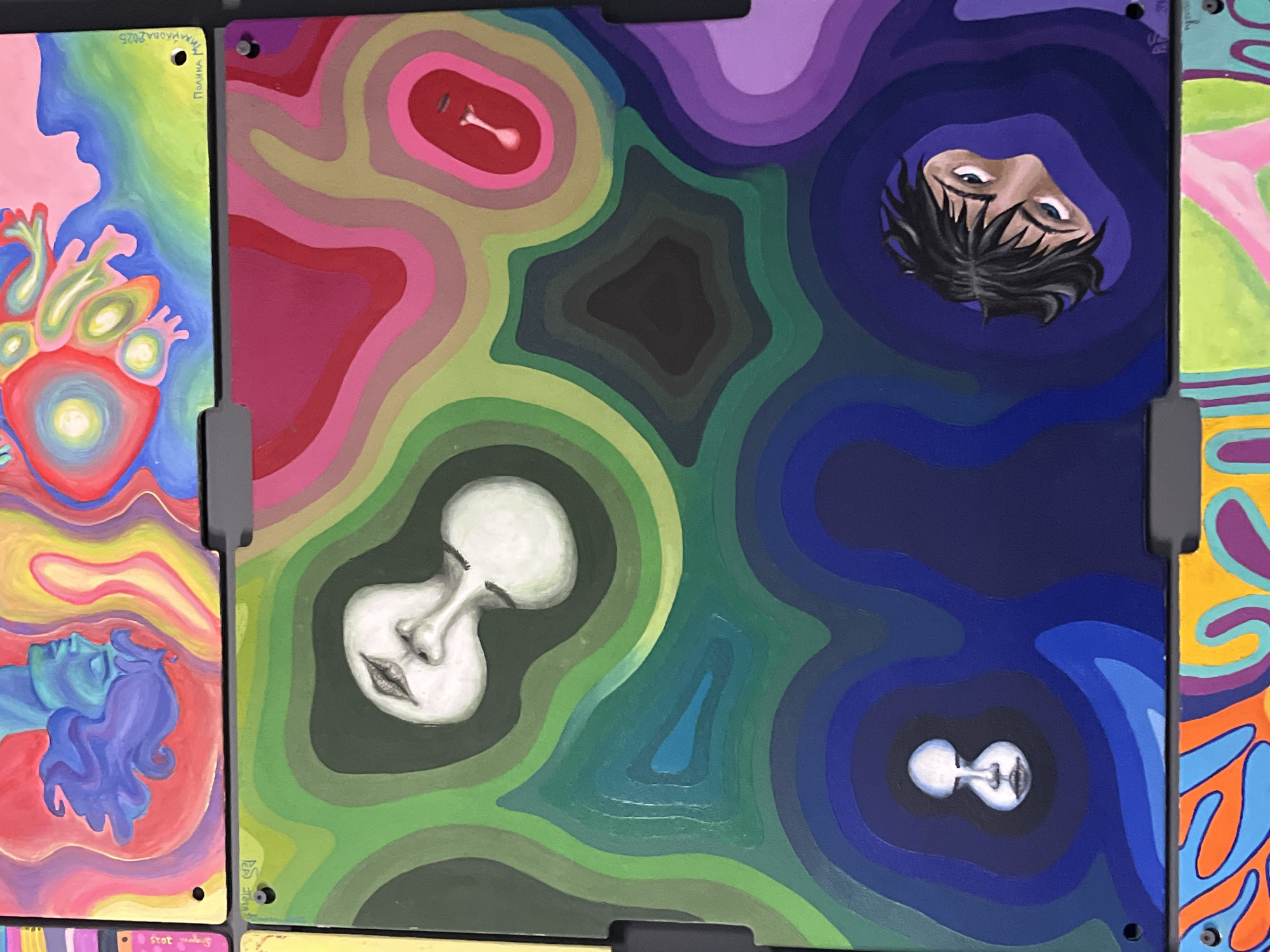 The image shows a collection of paintings, possibly part of an art installation or exhibition. The scene likely takes place indoors, as evidenced by the uniform background and the presentation of the paintings.
The paintings are mounted on panels, seemingly connected or displayed together. The panels are connected with gray hardware.
The visible paintings feature abstract and figurative elements:
- A painting on the left side of the image depicts a pink heart with a blue person figure below it.
- The central painting is a colorful abstract composition. It features concentric lines forming shapes of red, purple, green and blue and abstract faces.
- Another visible painting has a face, upside down, on the right side of the painting.
- A painting is partially seen on the right side of the image, with abstract shapes and a color scheme of yellow, orange, blue and purple.
The environment suggests an art gallery or public art space. The city of Sofia, Bulgaria, cannot be confirmed.