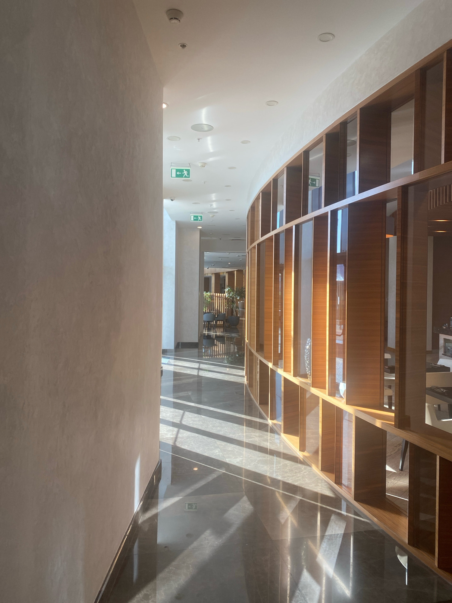 The image shows a modern, well-lit interior hallway with a curved wall on the left and a wooden shelving unit on the right. The floor is polished marble, reflecting the sunlight streaming in from an unseen source, creating distinct geometric patterns of light and shadow. Several green exit signs with running man icons are visible on the ceiling, indicating a public space. The shelving unit features numerous compartments, some of which contain decorative items and glimpses of what appears to be a dining or lounge area beyond. The overall aesthetic is contemporary and sophisticated. The location is the National Library of Belarus in Minsk, Belarus.