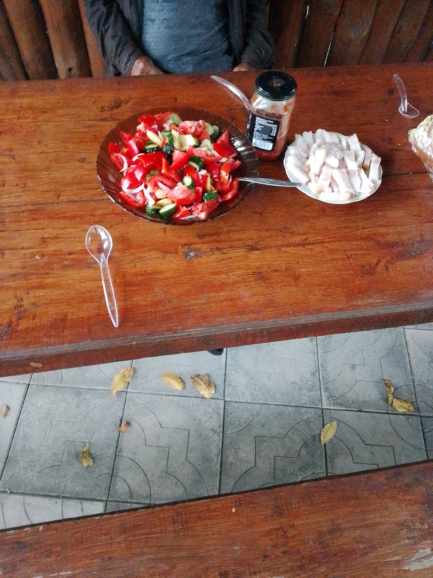The image depicts a wooden table with food and a person sitting in the background. On the table, there is a brown glass jar containing a red substance, likely a condiment or sauce. Next to the jar is a white plate filled with sliced white food, possibly pork fat or bacon. A salad made of chopped tomatoes, cucumbers, onions, and peppers is served on a dark brown plate. Two clear plastic spoons are also on the table, one near the salad and another to the right. The person in the background is wearing a gray long-sleeved shirt. The table appears to be outdoors, with a tiled floor visible below. The scene is consistent with a casual meal or gathering in Myrhorod, Ukraine.