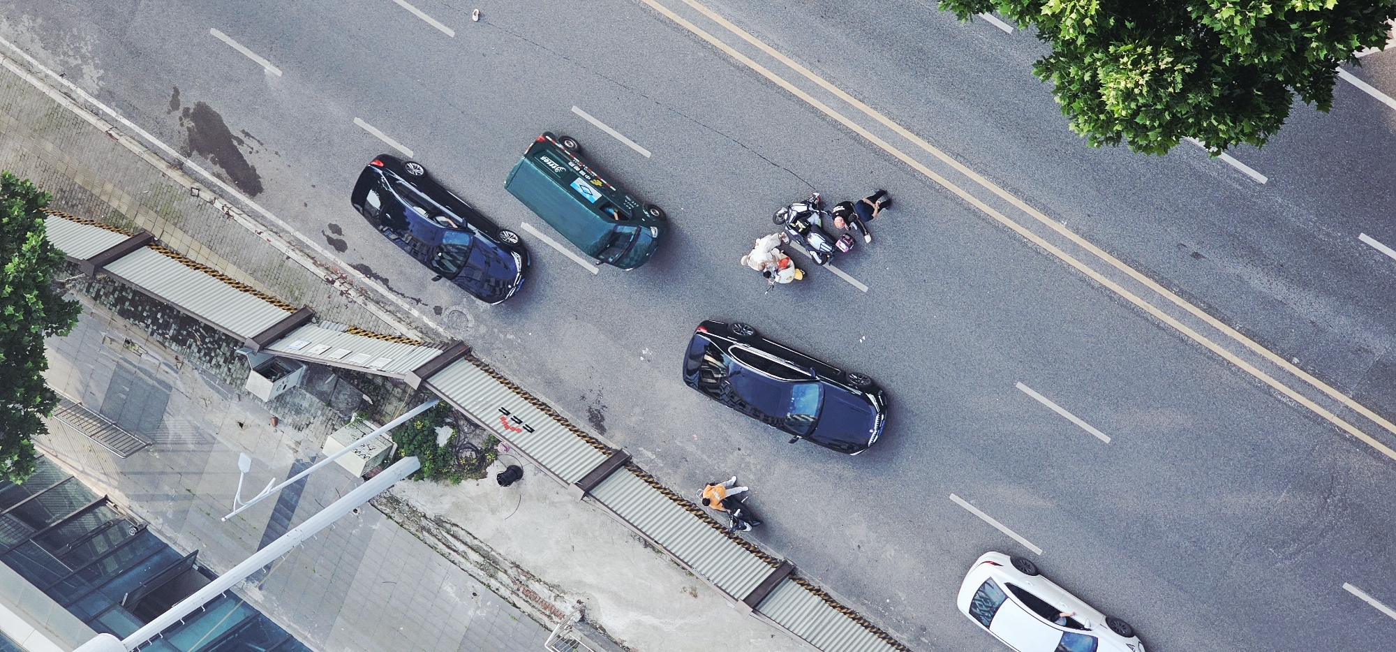 The image depicts an aerial view of a street in Jiulongpo District, China. Five cars are visible: two dark-colored sedans, a green van, and a white sedan. A group of people are gathered around a motorcycle in the middle of the street. Another person is near a parked car. A pedestrian bridge runs along the side of the street, adjacent to a building. Trees are visible on both sides of the street. Road markings and lane dividers are painted on the asphalt.