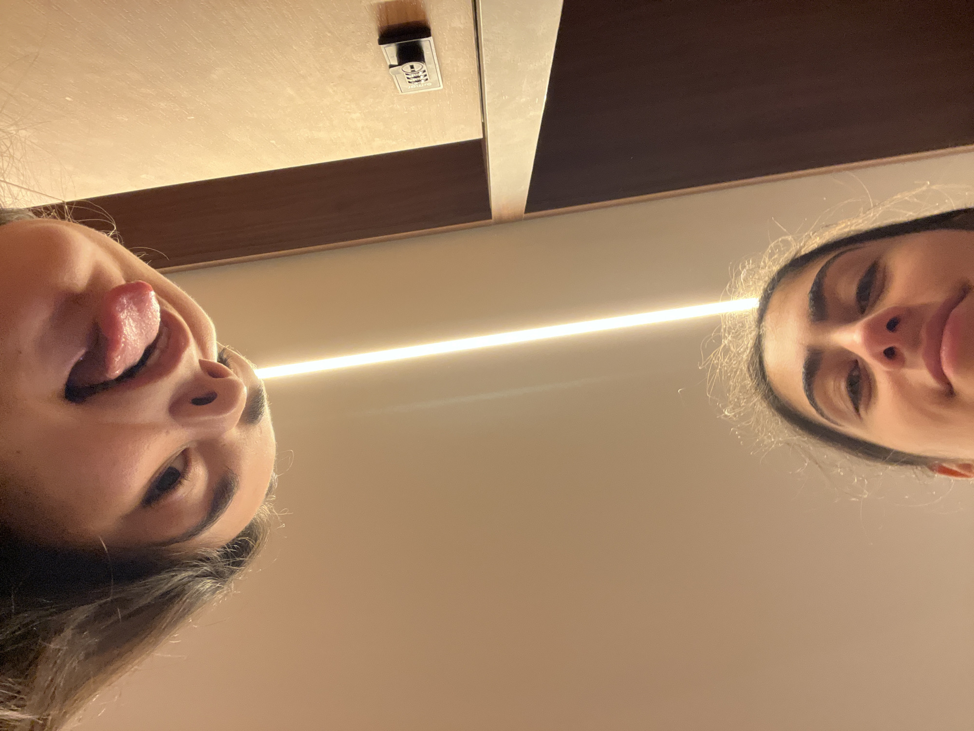 This is a low-angle, indoor photograph featuring the faces of two young women. The woman in the foreground, positioned at the bottom of the frame, is looking upwards towards the camera with a slight smile. Her facial features, including her eyebrows, eyes, nose, and lips, are visible. Her hair is slightly messy. The woman in the background, at the top of the frame, is also looking down towards the camera. Her tongue is sticking out, and her eyes are partially closed.
The environment appears to be a room with a light-colored ceiling illuminated by a vertical strip of light. To the right of the frame, a dark wooden surface is visible, with a small black combination lock attached to it. The overall lighting suggests an interior setting, possibly a locker or closet area. There is no indication of the city of Paris, France, or any other specific location.