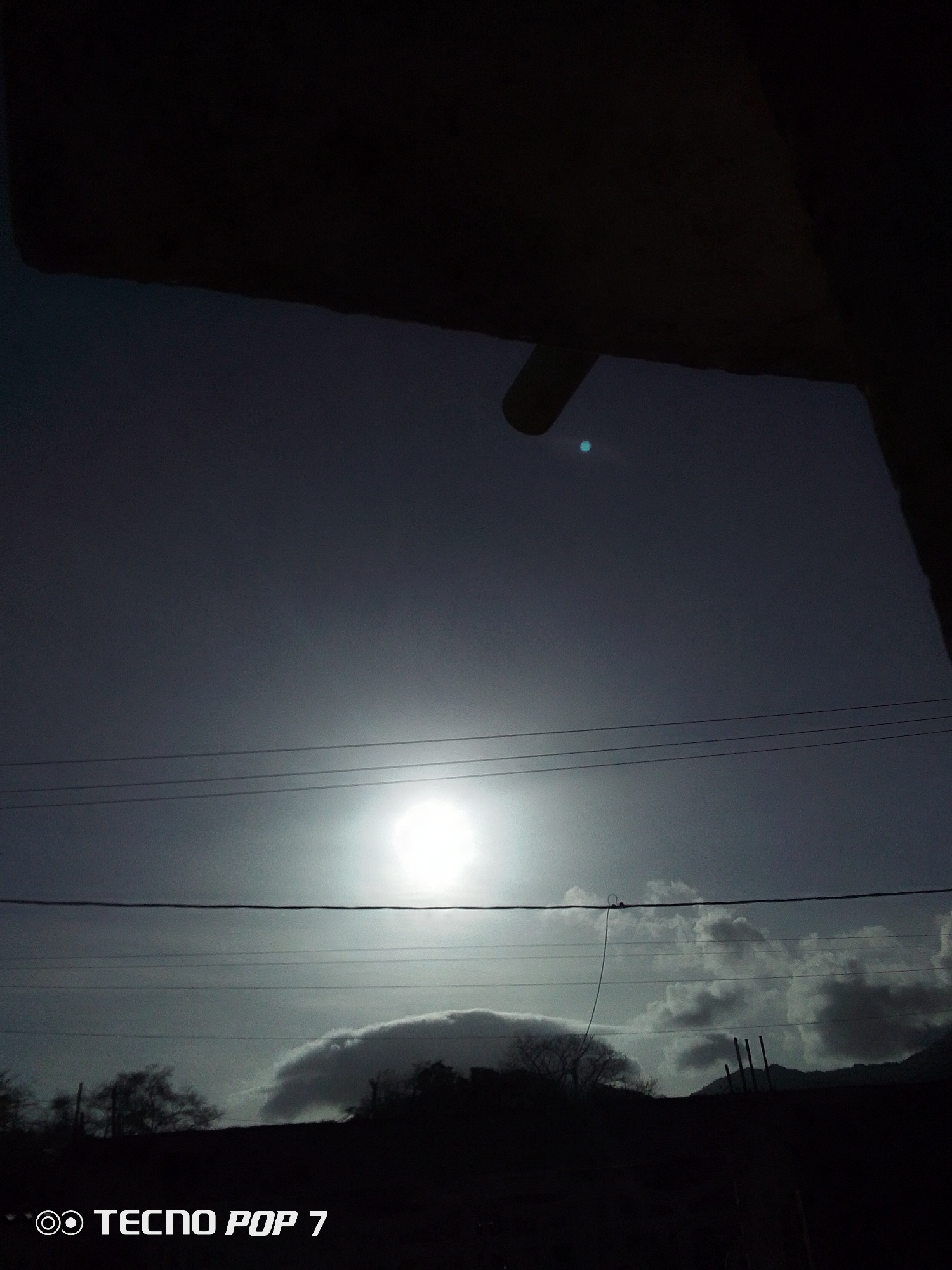 The image shows an outdoor scene taken during daylight in Altagracia, Venezuela. The sun is visible and partially obscured by clouds. A few power lines run horizontally across the middle of the frame. The bottom portion shows silhouetted land with some trees and cloud formations. The top of the frame is partially covered by an unidentifiable dark, possibly overhanging, structure. At the bottom of the frame, the watermark for "TECNO POP 7" is seen, indicating the phone model used to take the picture.