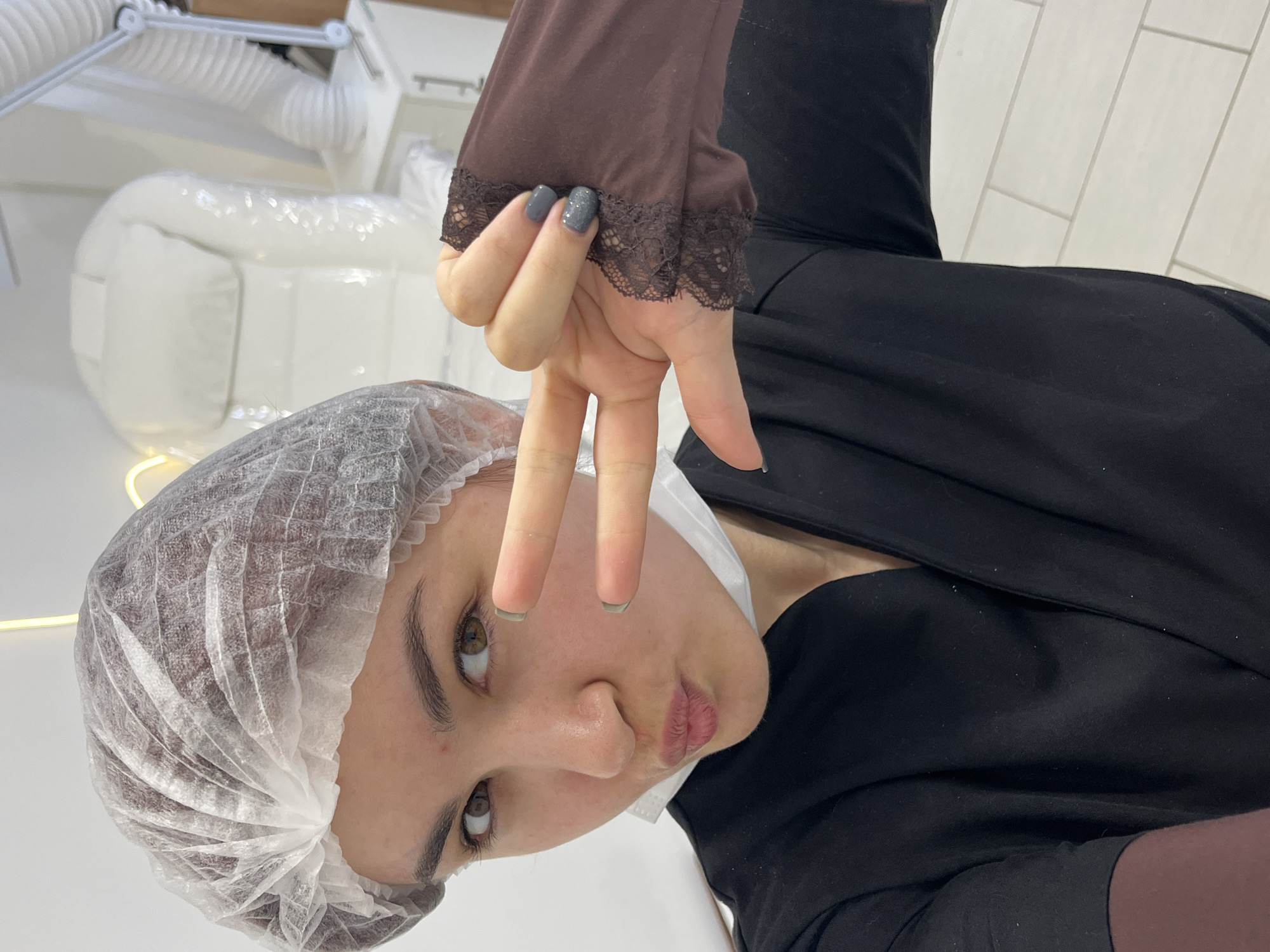 A female subject, located in Neftekamsk, Russia, is captured in a close-up, chest-up portrait within an indoor professional setting, consistent with a beauty or medical treatment room. She wears a translucent white disposable bouffant cap, a white surgical mask positioned under her chin, and a black uniform top layered over a dark brown long-sleeved garment with brown lace cuffs. Her right hand is raised, displaying a "peace" gesture with extended index and middle fingers; her fingernails are polished in dark grey, with the middle finger featuring glitter. Her expression is a pout with wide-open brown eyes, looking directly at the camera. The background features white walls, a white tiled floor, a white reclining treatment chair covered in clear plastic sheeting, a white cabinet, an articulated white lamp, and a white corrugated ventilation tube. Warm-toned linear and curved LED light fixtures are visible on the wall.