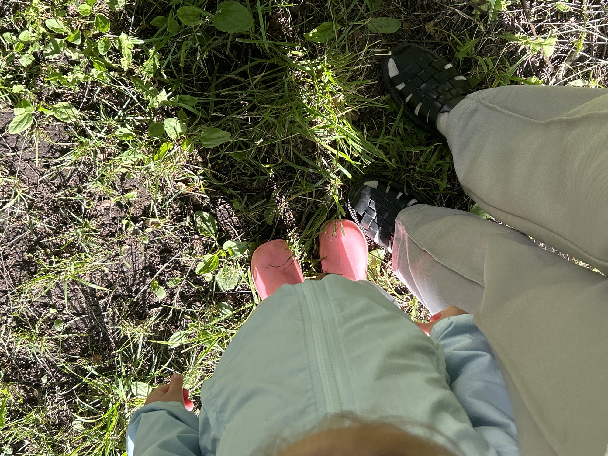 The image shows a top-down view of two people's legs and feet standing on grass and dirt. One person is wearing black woven sandals and light-colored pants. The other person, likely a child, is wearing pink rain boots and a light blue hooded jacket. The grass appears to be growing in patches, with some areas of bare dirt. The lighting suggests it is daytime, with shadows cast by the sun. The overall scene appears to be outdoors, possibly in a park or backyard. There is no visible text in the image. The image is a close-up of the feet and lower legs, with the majority of the frame occupied by the ground and foliage. The focus is on the footwear and the natural environment.