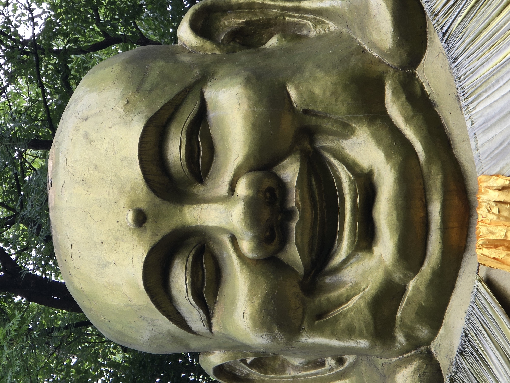 A close-up, full-frame shot shows the gilded face of a large statue of a laughing Buddha. The statue is golden yellow and has a very round face with closed eyes, a wide smile, and prominent cheeks. The statue is positioned outdoors, with green foliage and tree trunks visible in the background. The bottom of the statue is draped with fabric, some of which is golden yellow. The image appears to be of a statue located in the Xihu District of China.