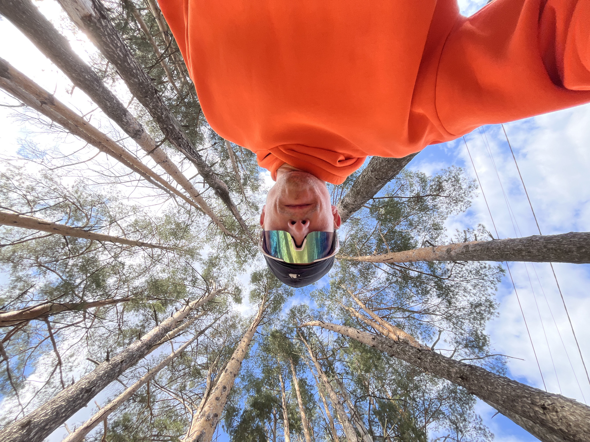 A unique low-angle, wide-angle shot captures an inverted person against a backdrop of tall pine trees and a partly cloudy sky. The main subject, a person, is seen from a perspective looking directly up at them. They are wearing a vibrant orange hooded sweatshirt, a dark cap, and large, reflective sports sunglasses that show multi-colored hues of green, blue, and purple. Their face, visible from the chin up, appears at the bottom center of the frame, while their orange hoodie fills much of the upper portion, creating an illusion of being suspended upside down.
The background features numerous slender pine tree trunks extending upwards, with some branches and green needles visible against the bright sky. Patches of clear blue sky with fluffy white clouds are interspersed between the tree canopy, indicating a bright daytime setting. Thin lines, possibly ropes or wires, are visible against the sky on the right side of the image, suggesting an adventure park or outdoor activity. The photograph provides an immersive view into a forest environment, consistent with a natural setting like Chaikovsky, Russia. No text is visible in the image.