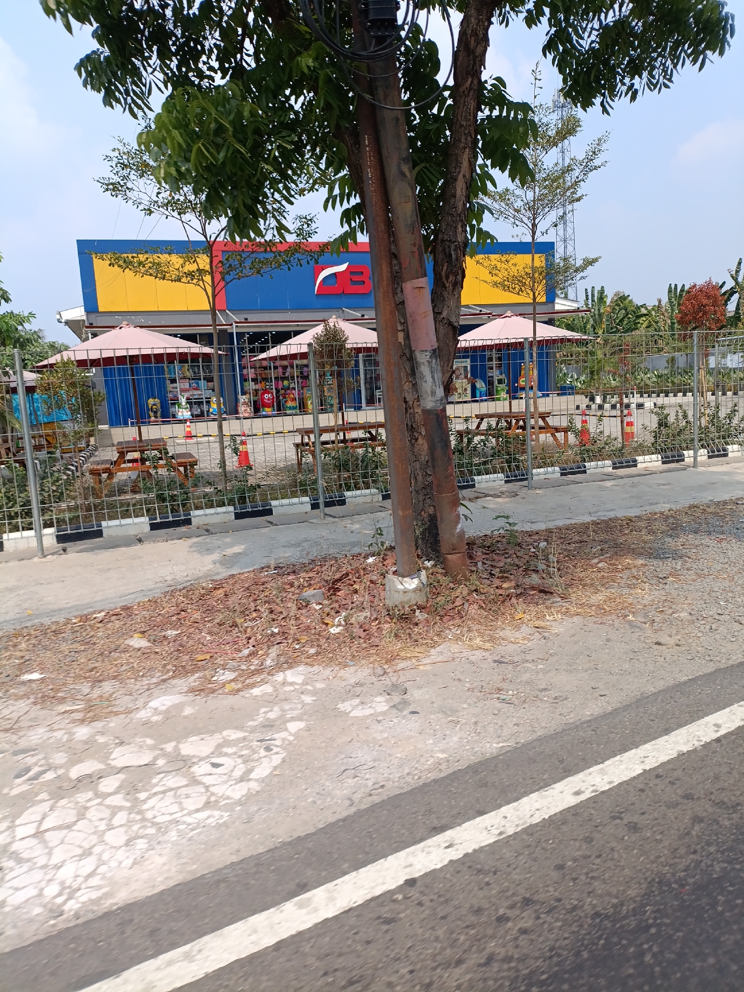 This is a photograph of a storefront with a blue and yellow facade, likely a retail establishment, located in Cariu, Indonesia. The building features large windows and a prominent logo that appears to be "CB". In front of the building, there are several picnic tables and red umbrellas providing shade. A wire fence encloses the area in front of the building, separating it from the street. A prominent utility pole stands in the foreground, partially obscuring the view of the building and surrounding vegetation. The ground in front of the fence is covered with dry leaves and debris. A road with a white line marking is visible in the bottom right corner of the image. The sky is clear and blue, indicating a sunny day. No people are clearly visible in the frame.