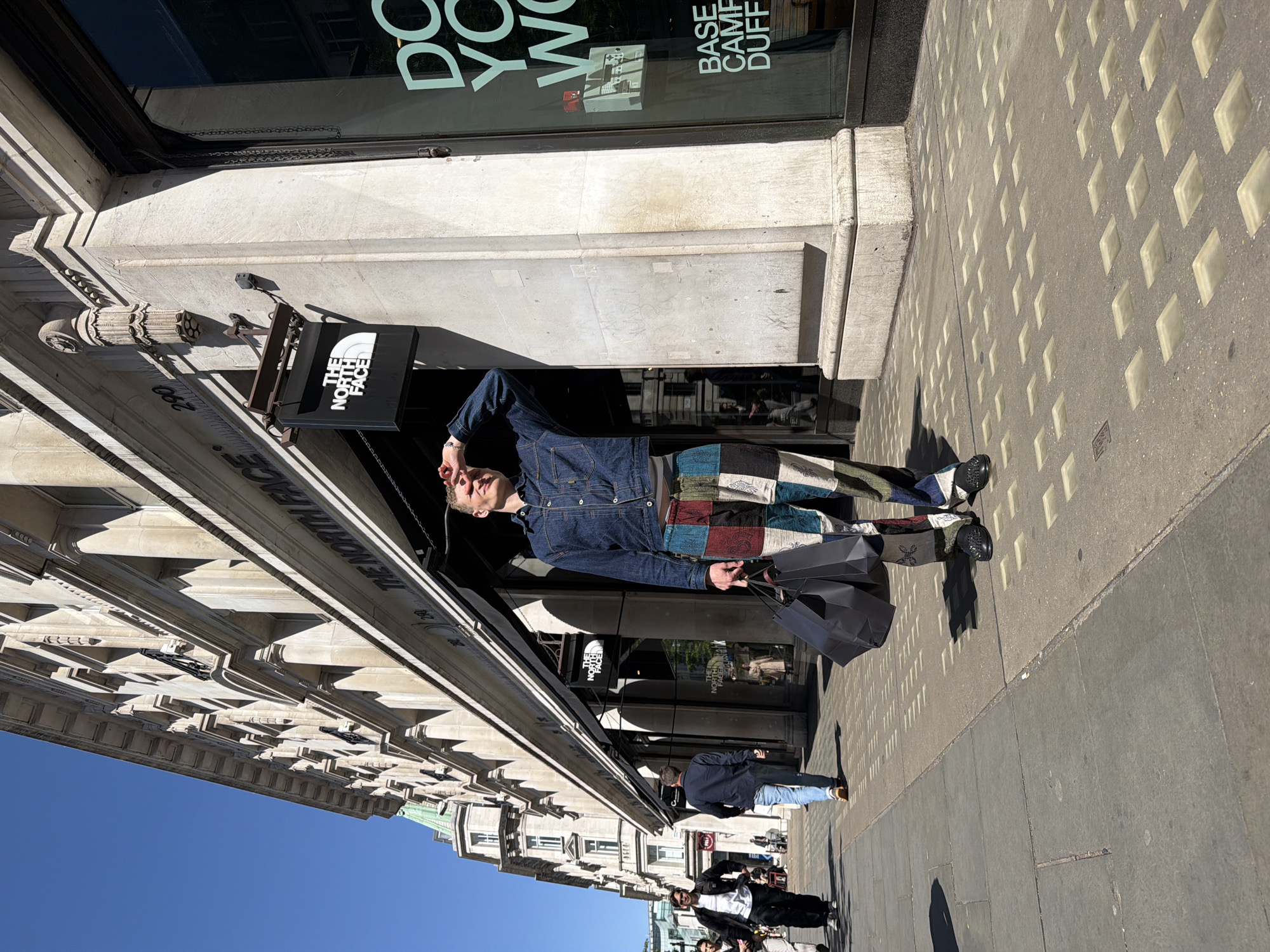 A man in a denim jacket and patchwork pants stands in front of The North Face store on Regent Street in London. He is holding shopping bags. The sky is blue. Other people walk along the sidewalk.
