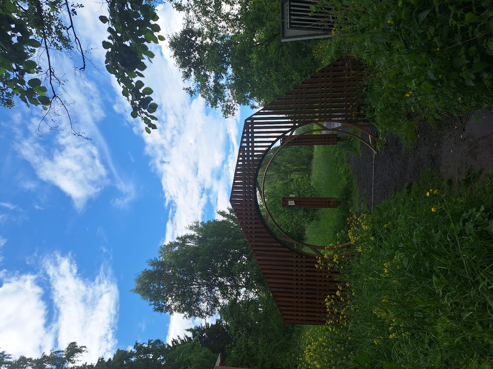 The image depicts an outdoor scene in the city of Sim, Russia. In the foreground, a gravel path leads towards a large, circular wooden archway. The archway is constructed of vertical wooden slats and appears to be part of an entrance or gateway. Surrounding the path and archway is lush green vegetation, with tall grasses and scattered yellow wildflowers.
Beyond the archway, the path continues into a wooded area with numerous trees and foliage. The sky above is a bright blue, dotted with fluffy white clouds. To the right of the archway, a metallic structure with vertical bars is partially visible, suggesting it might be part of a fence or enclosure. The overall impression is of a natural, park-like setting, possibly an entrance to a trail or a recreational area.