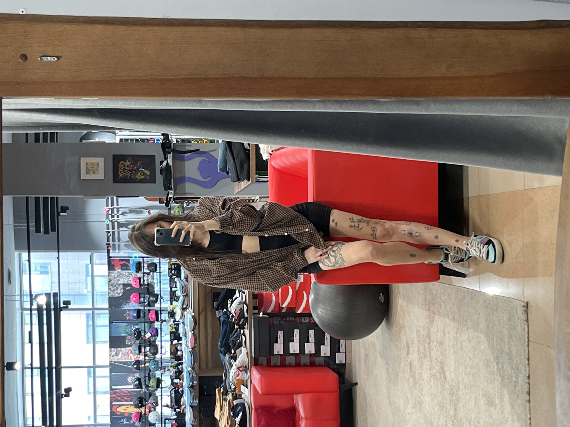 A woman is visible in a mirrored reflection. She is sitting on a red chair, resting her leg on a gray exercise ball. The woman is taking a photo with her cell phone. Her legs are visible, and they have multiple tattoos. She is wearing a black top and a brown checkered jacket. Shelves of snowboards and shoes are displayed in the background. The reflection is taking place in a store that sells sporting goods. The location is likely in Krasnoyarsk, Russia.