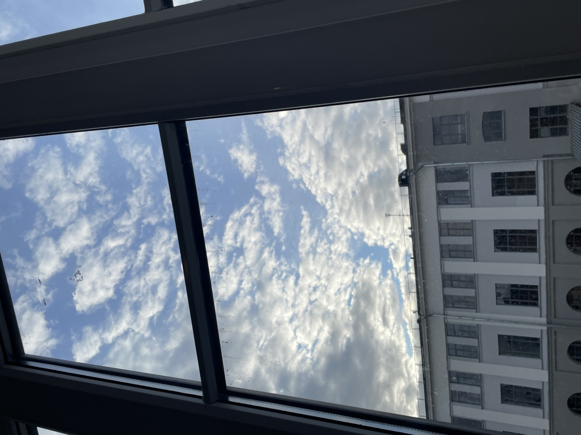 The image presents an upward view through a glass ceiling or skylight, featuring both the sky and a reflected building. The central and left portions of the image are dominated by a bright blue sky filled with scattered white, fluffy clouds. Numerous small specks of dust or dirt, along with some streaks, are visible on the surface of the glass, particularly evident against the sky.
To the right, reflected in the glass, is a multi-story building. This structure is light-colored, appearing white or light grey, and features a repetitive architectural design with various window shapes. Rectangular windows are most common, but some windows have arched tops, and at least one oval window is visible towards the bottom right of the reflection. The building's style suggests a classic or historic design, typical of urban architecture.
The scene depicts daytime with fair, cloudy weather. There are no people visible, nor is there any discernible text. The image provides a static view, combining an atmospheric sky with an architectural reflection, taken from a low vantage point looking upwards through a glass structure. The location is Saint Petersburg, Russia.