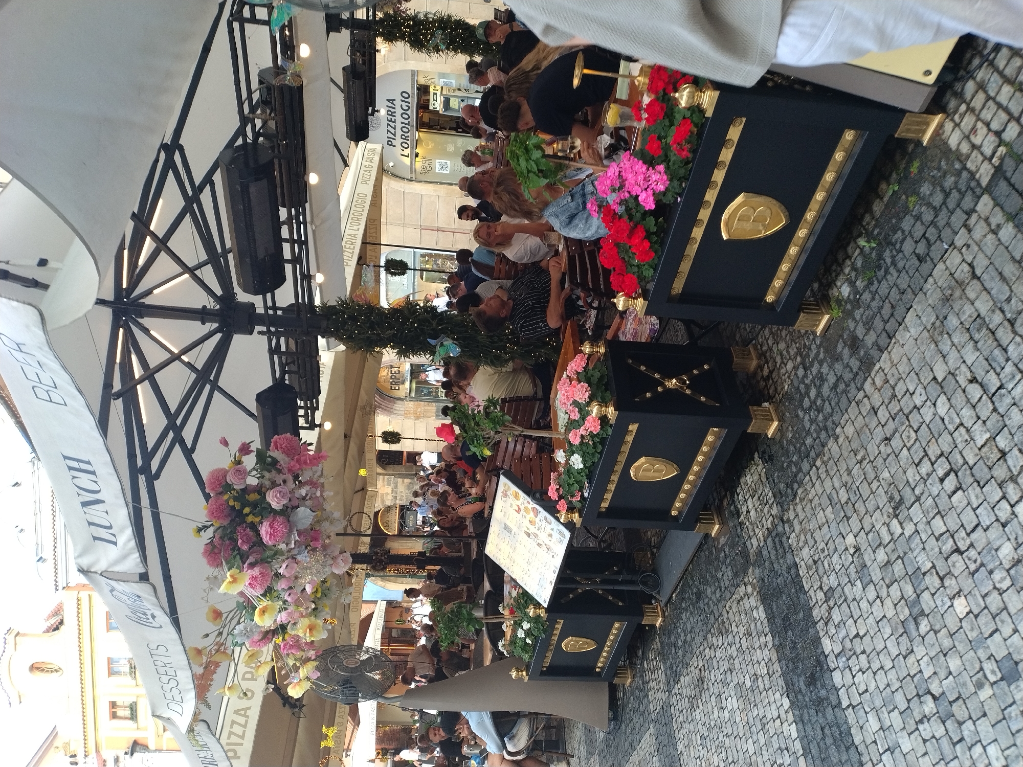 This image captures an outdoor seating area of a restaurant, likely in Prague, Czechia, judging by the architecture visible in the background. The scene is set under a large white umbrella, adorned with the words "LUNCH" and "BEER". A vibrant floral arrangement hangs from the umbrella's structure, adding a splash of color to the scene.
In the foreground, black planters with gold accents and a stylized "B" emblem are placed along the perimeter. These planters are filled with colorful flowers, predominantly pink and red, with some white blooms. A menu board stands on a stand between two of these planters.
Numerous people are seated at tables, enjoying meals and drinks. Some are casually dressed, while others appear to be in more formal attire. The atmosphere suggests a lively dining experience. The ground is paved with cobblestones, typical of historic European cities. Restaurant awnings and signage, including "PIZZA L'OROLOGIO", are visible in the middle ground, indicating the establishment's offerings. There's also a prominent sign for "PIZZA & PASTA". Various decorative elements, such as string lights and small potted trees, contribute to the ambiance. A large industrial fan is also visible to the left of the image.