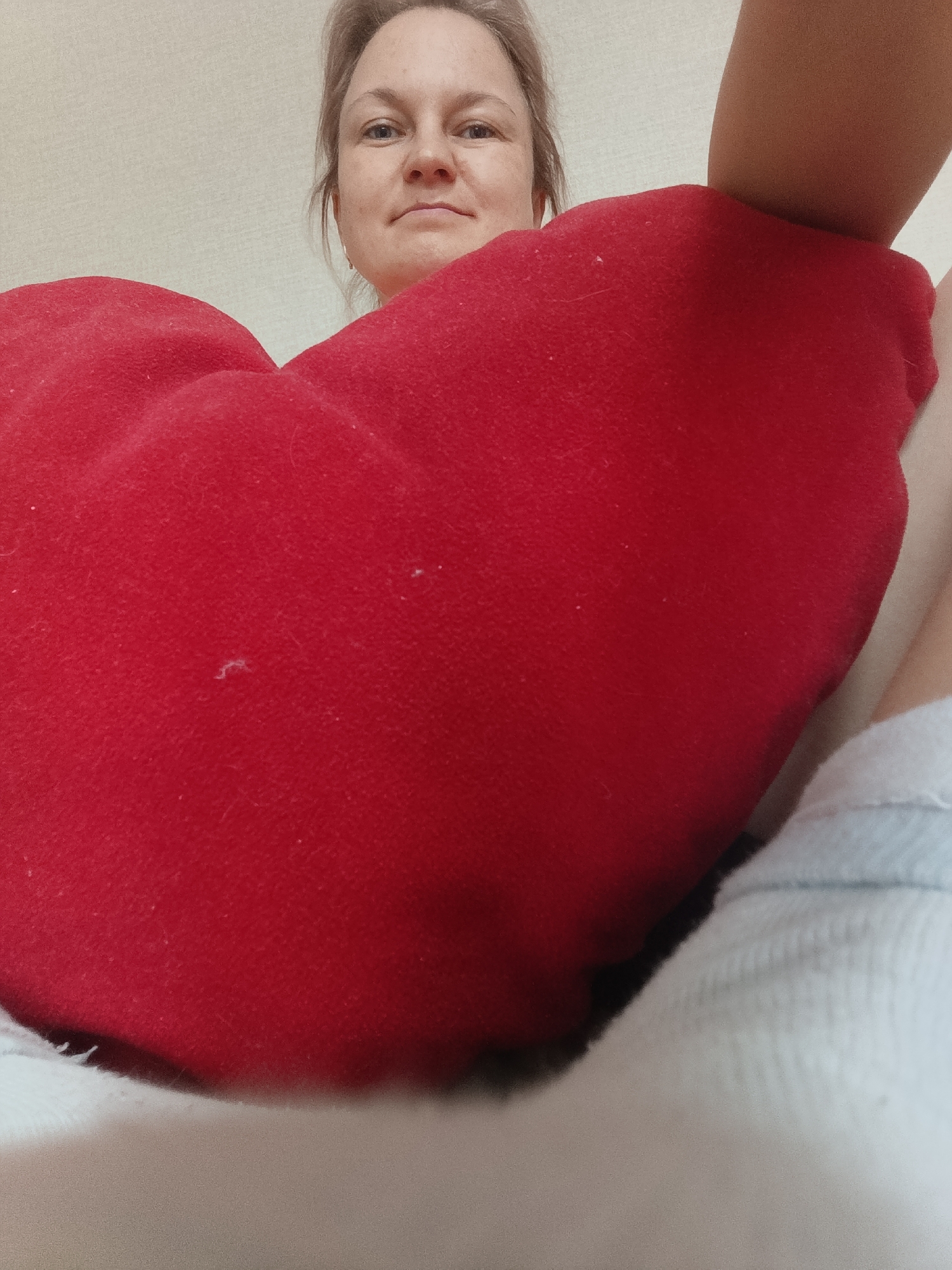 A woman is holding a large, red, heart-shaped pillow. The woman's face is visible above the pillow; she has fair skin and her hair is pulled back. A person's arm is partially visible on the upper right of the frame. She is sitting in a bed with a light blue and white blanket or sheet. The background is a plain, light-colored wall. This image does not contain any clues as to the city of Марковский, Russia.