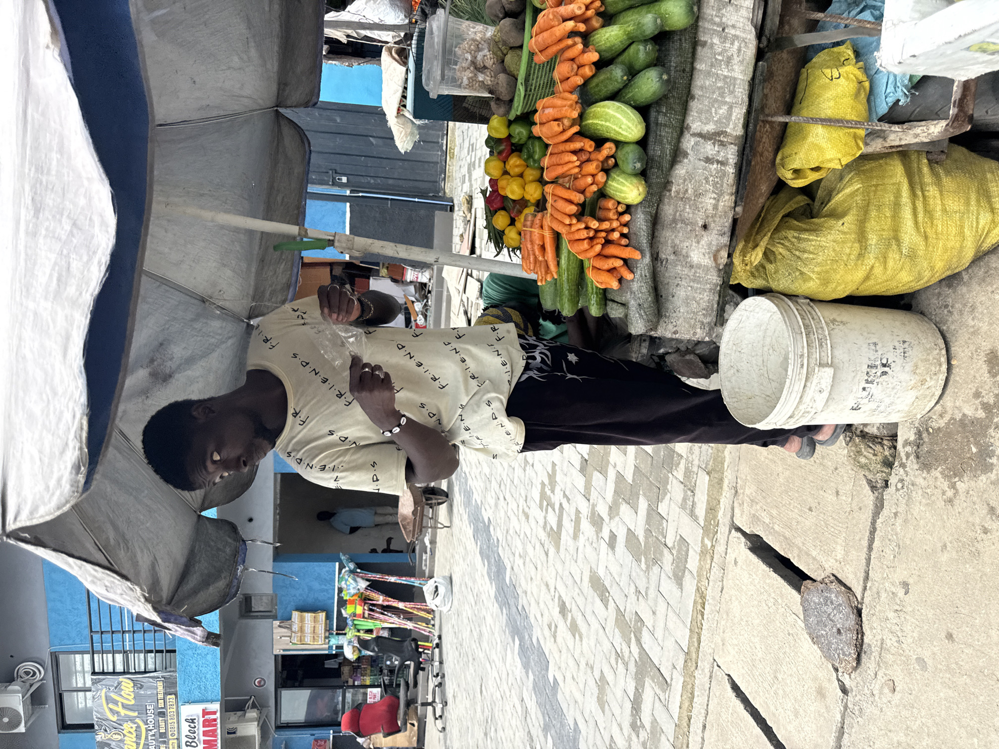 A man stands under a weathered white umbrella at a street market, holding a plastic bag. He is wearing a light-colored t-shirt with repeated white lettering and dark pants. In front of him is a wooden cart laden with fresh produce, including carrots, cucumbers, bell peppers, and other vegetables. To his left, a white bucket rests on the ground. The market appears to be located in Ado, Nigeria, indicated by a sign in the background that reads "BLOCH MART" and appears to be in English, a common language in Nigeria. The ground is paved with interlocking bricks, and the buildings in the background have blue facades. The scene suggests a typical day at a local food market.