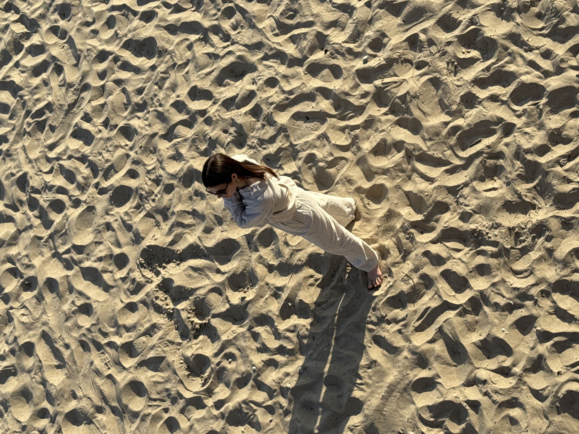 An aerial, high-angle shot captures a person from the chest down, standing on a sandy beach. The individual is wearing light-colored, loose-fitting clothing, possibly a jumpsuit, and sunglasses. Their arms are crossed, and they are looking off to the left. Bare feet are visible, pressing into the sand. The sand itself is textured with numerous footprints and ripples, indicative of a beach environment. The lighting suggests it is daytime, with shadows cast by the person and the sand formations. The scene does not provide any specific visual cues that would allow for definitive identification of its location within Tallinn, Estonia.