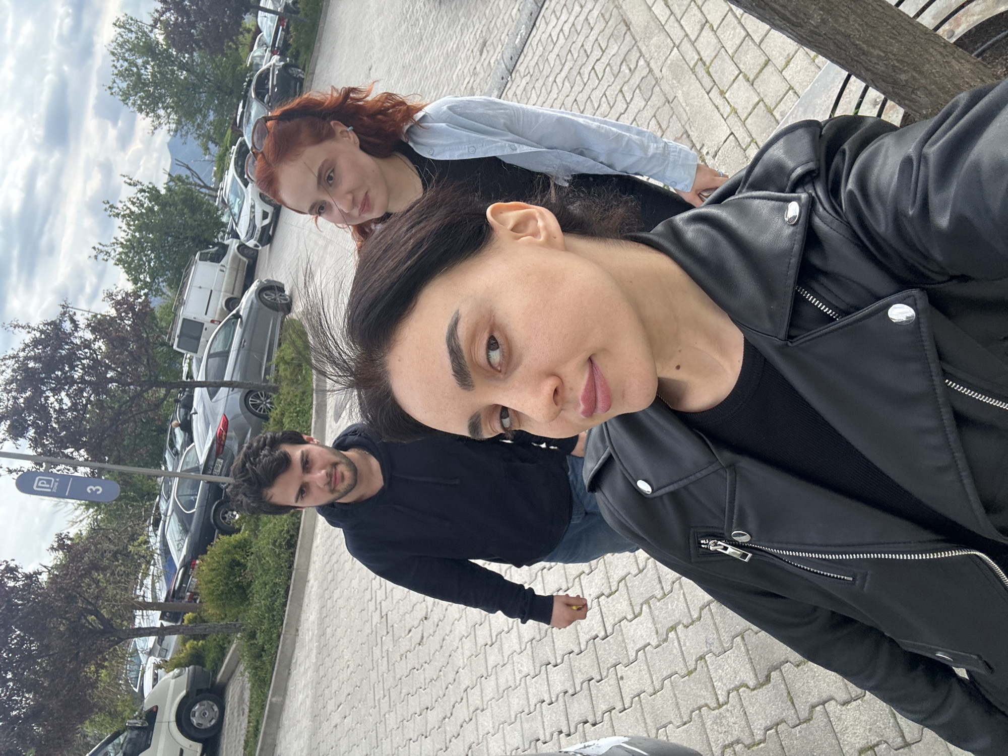 The image depicts three individuals in what appears to be an outdoor parking area under an overcast sky. The photograph is taken from a high, angled perspective, suggesting a selfie.
In the immediate foreground, a woman with dark hair wears a black leather jacket over a dark top. She looks directly at the camera with a neutral expression, her lips slightly parted. Her right ear and a mole on her neck are visible.
Behind her and to the left, a man with dark hair and a beard is dressed in a black hooded sweatshirt and jeans. He holds a small yellow item, possibly a fruit or snack, in his right hand and looks towards the camera with a calm expression.
Further back and to the right, another woman with reddish-brown hair is visible. She wears a light blue button-up shirt over a dark top and has sunglasses perched on her head. She offers a subtle, gentle smile towards the camera.
The background features a line of parked cars, including a white van, along a paved surface made of light-colored interlocking pavers. Trees and green bushes are interspersed along the parking lot. A section of a dark bench is visible on the right side of the frame. A blue parking sign with a white "P" symbol and the number "3" is prominently displayed on a pole among the vehicles. The lighting is soft and diffused, indicating daytime.