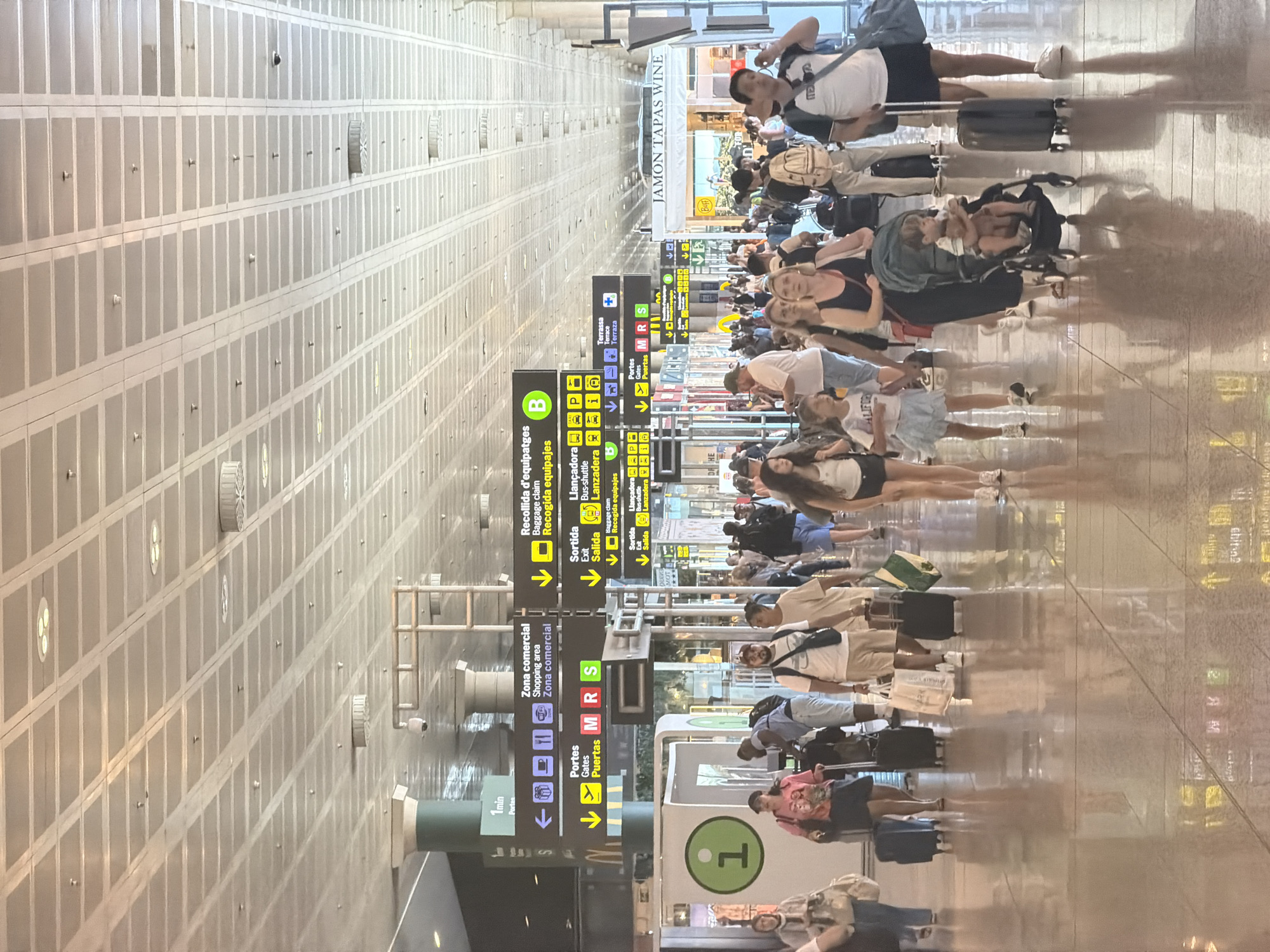 The image shows a bustling interior scene of a modern airport terminal, likely in El Prat de Llobregat, Spain, given the signage. A large number of people are walking in various directions, many with luggage, indicating travel. The environment features a high, tiled ceiling with recessed lighting, a polished reflective floor, and numerous directional signs in multiple languages. These signs provide information on commercial areas, shopping, baggage claim, gates, exits, bus shuttles, terraces, and other airport services. Visible are shops, informational kiosks, and structural elements like columns. The lighting is bright and even, suggesting an indoor environment. The focus is on the movement and activities of travelers within the airport concourse.